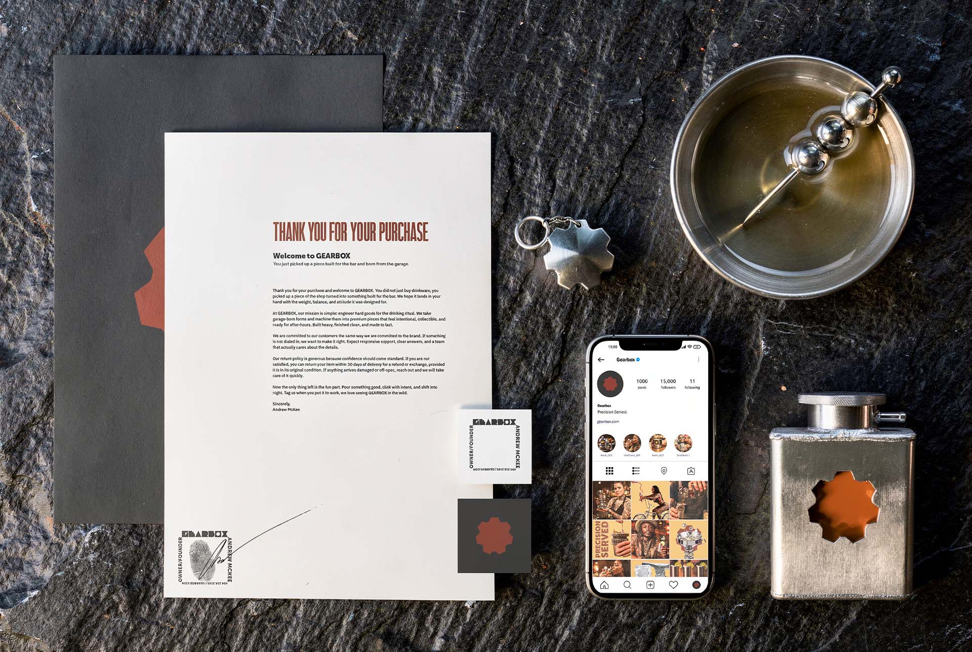 Gearbox stationery flat lay on slate with a welcome letter, cog keyring, business card, metal coupe, flask and the brand's Instagram profile on a phone