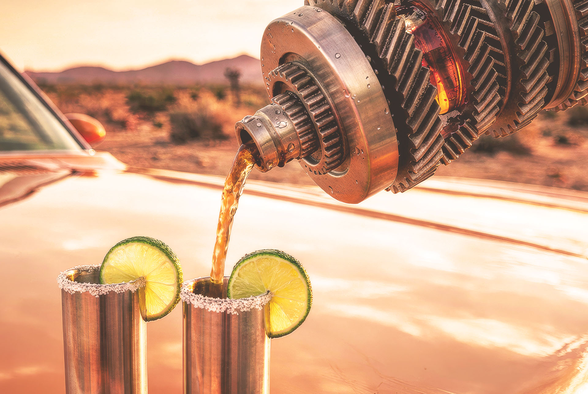 Gearbox transmission assembly pouring a cocktail into two salted rim metal tumblers with lime wheels, on a car hood in the desert at sunset