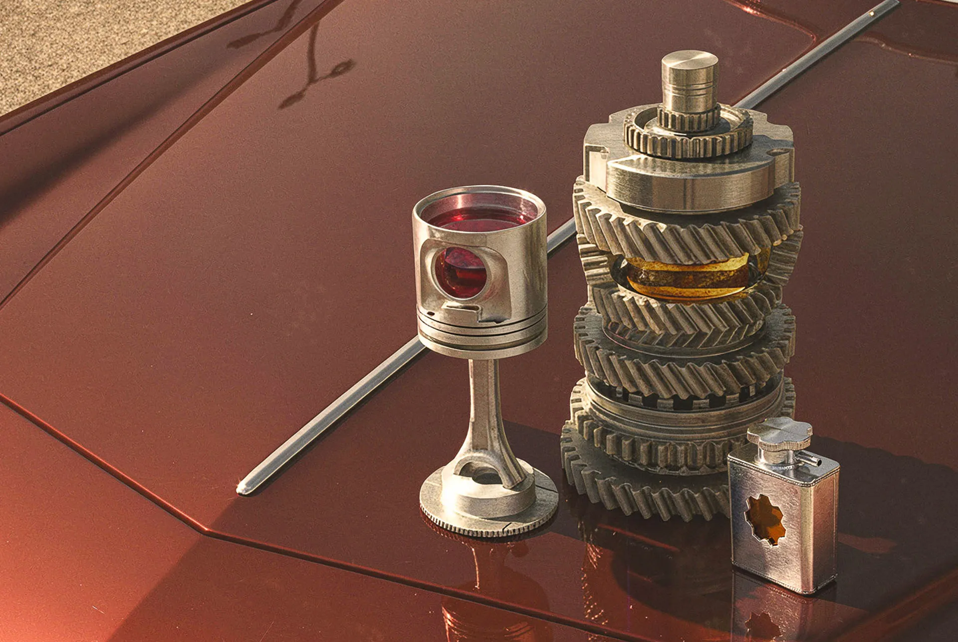 Gearbox piston stem wine glass filled with red wine beside a stacked gear shaker and a small hex flask on an oxblood car hood