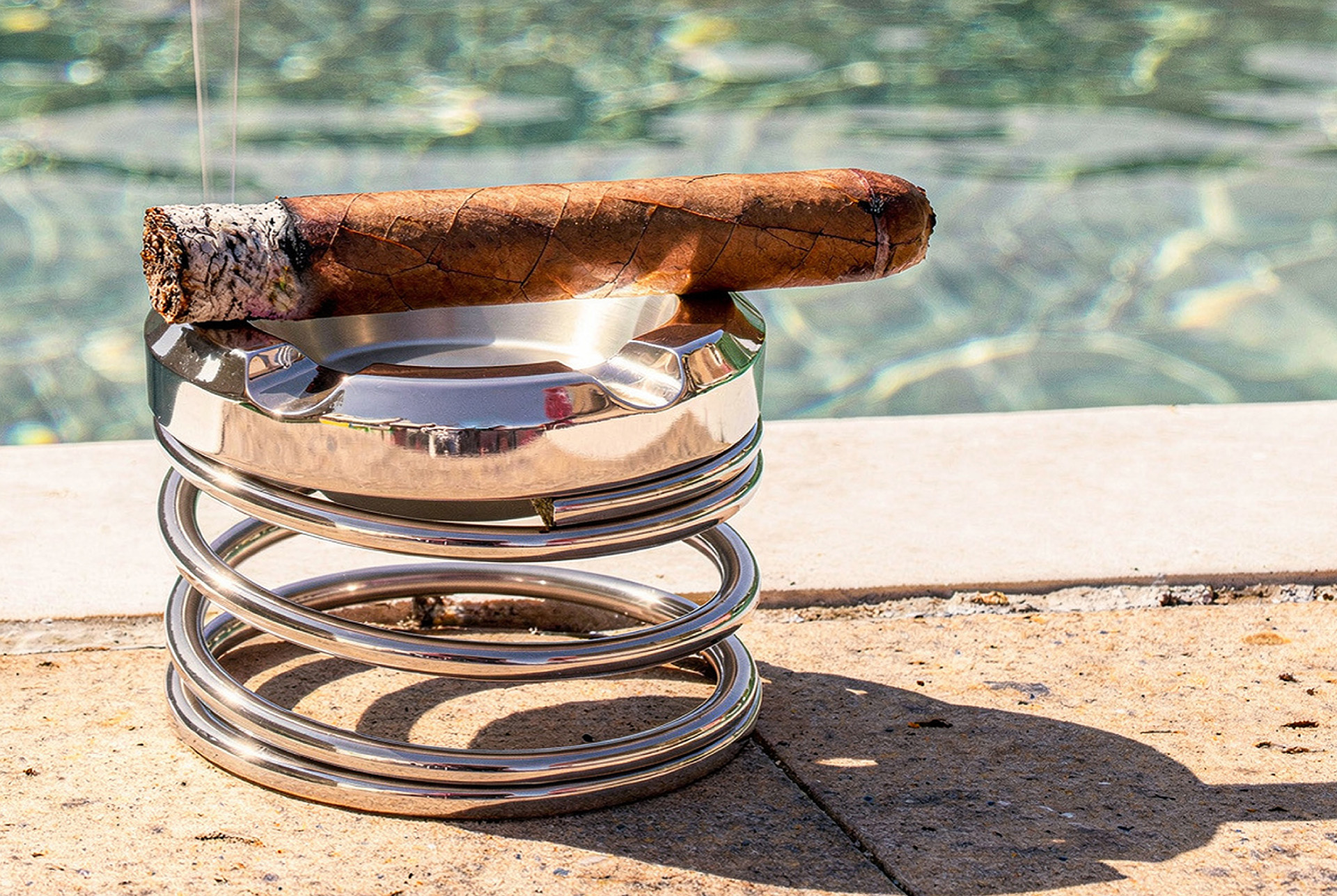 Gearbox coil spring ashtray in chrome holding a lit cigar, on pool coping with water behind it