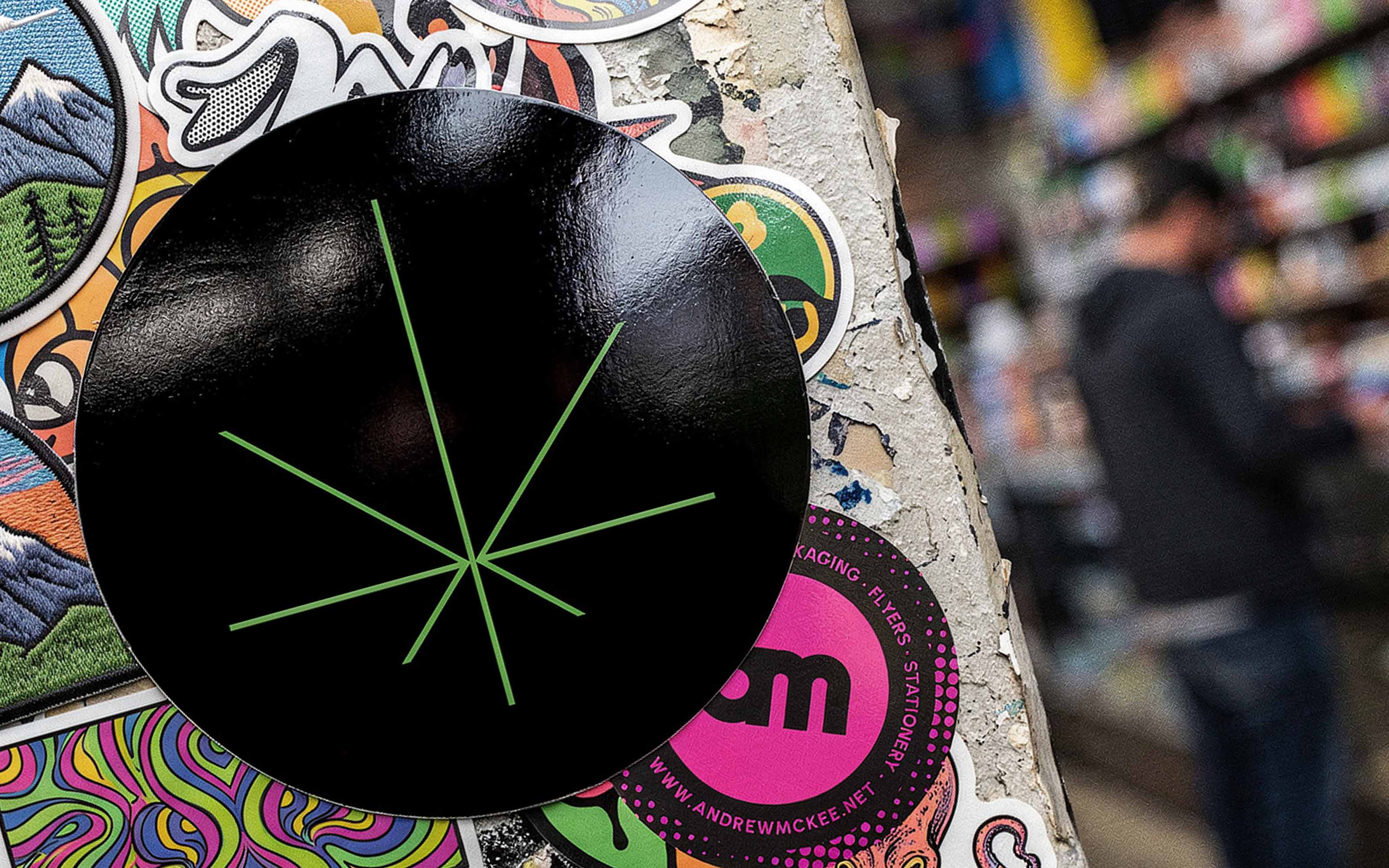 Black circular sticker with a green starburst mark, on a street pole layered with other stickers