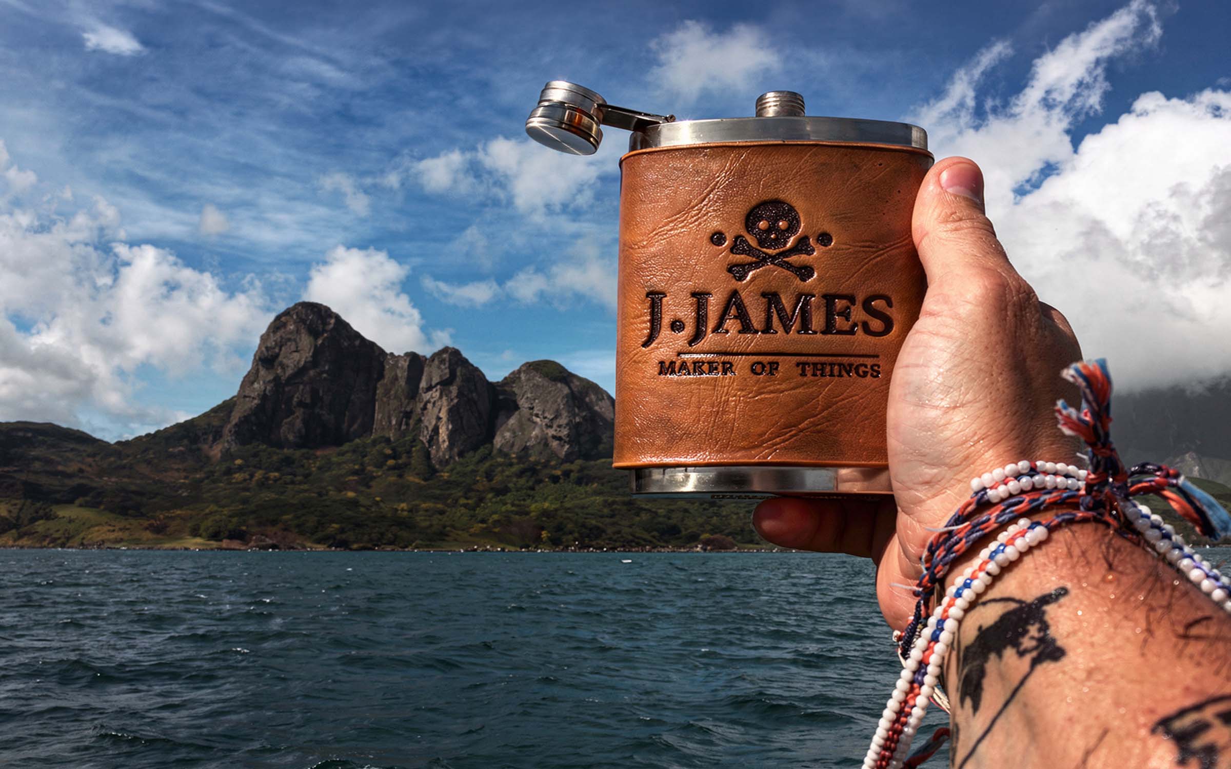 Hand holding the leather wrapped J. James flask above the water, with a volcanic peak behind it