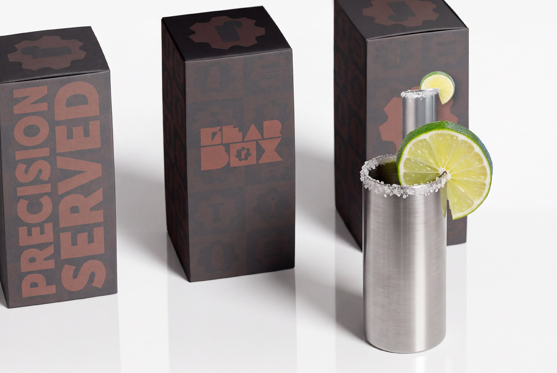 Three Gearbox boxes in the oxidized bronze gear pattern, one reading PRECISION SERVED, beside a brushed metal tumbler with a salted rim and lime