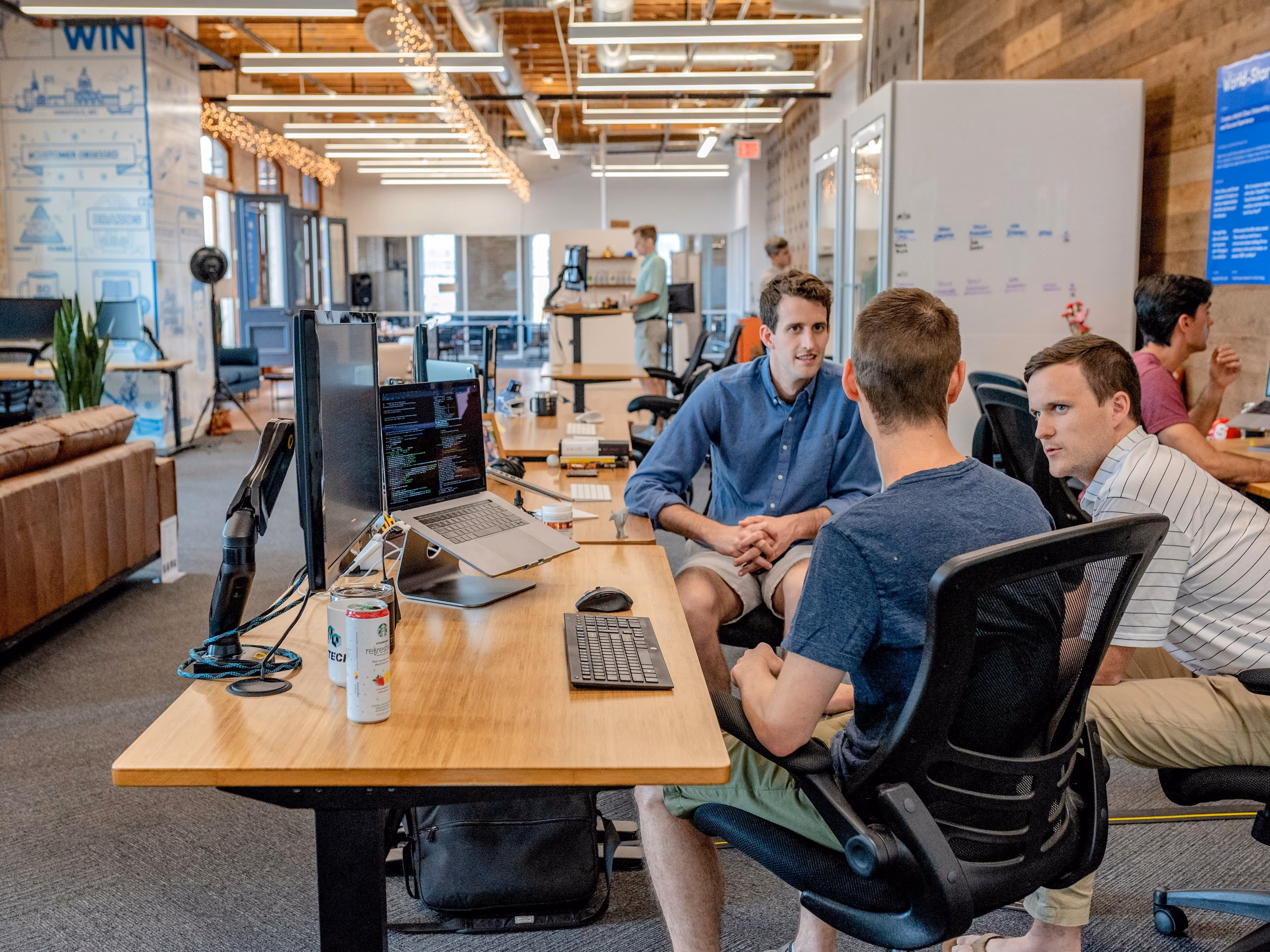 Three men in casual clothes having a discussion at an office desk with computer screens and laptops.