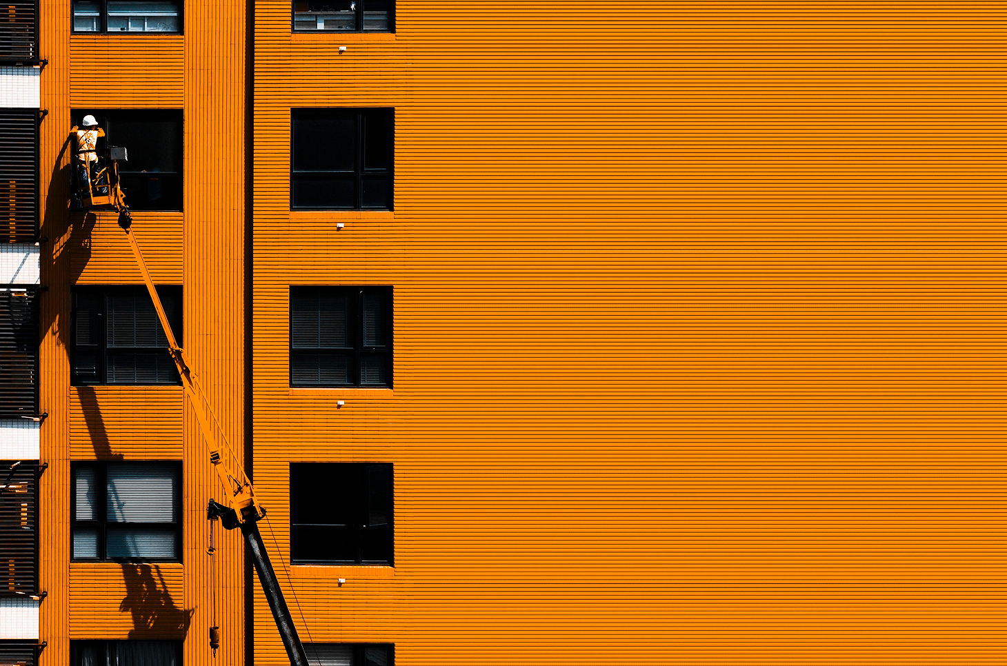 Construction worker wearing a white helmet on a yellow lift platform working on the orange exterior of a building with black-framed windows.