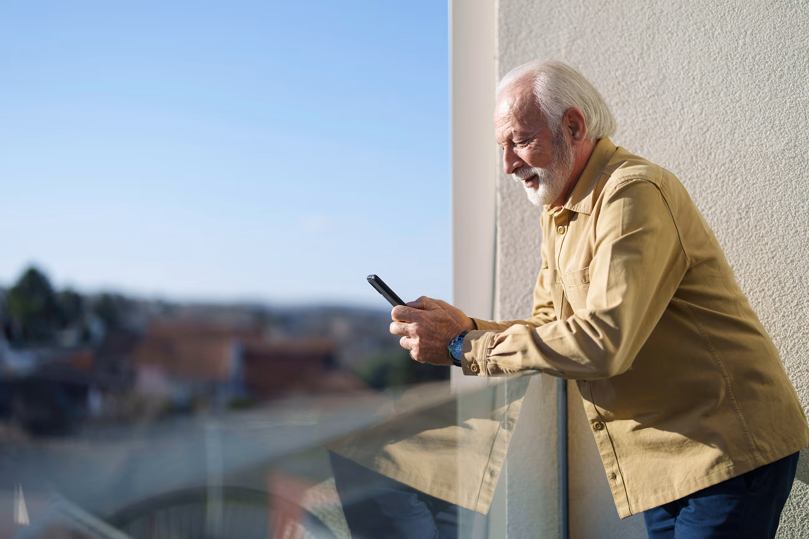 Older man leaning on balcony railing looking at smartphone with a slight smile.