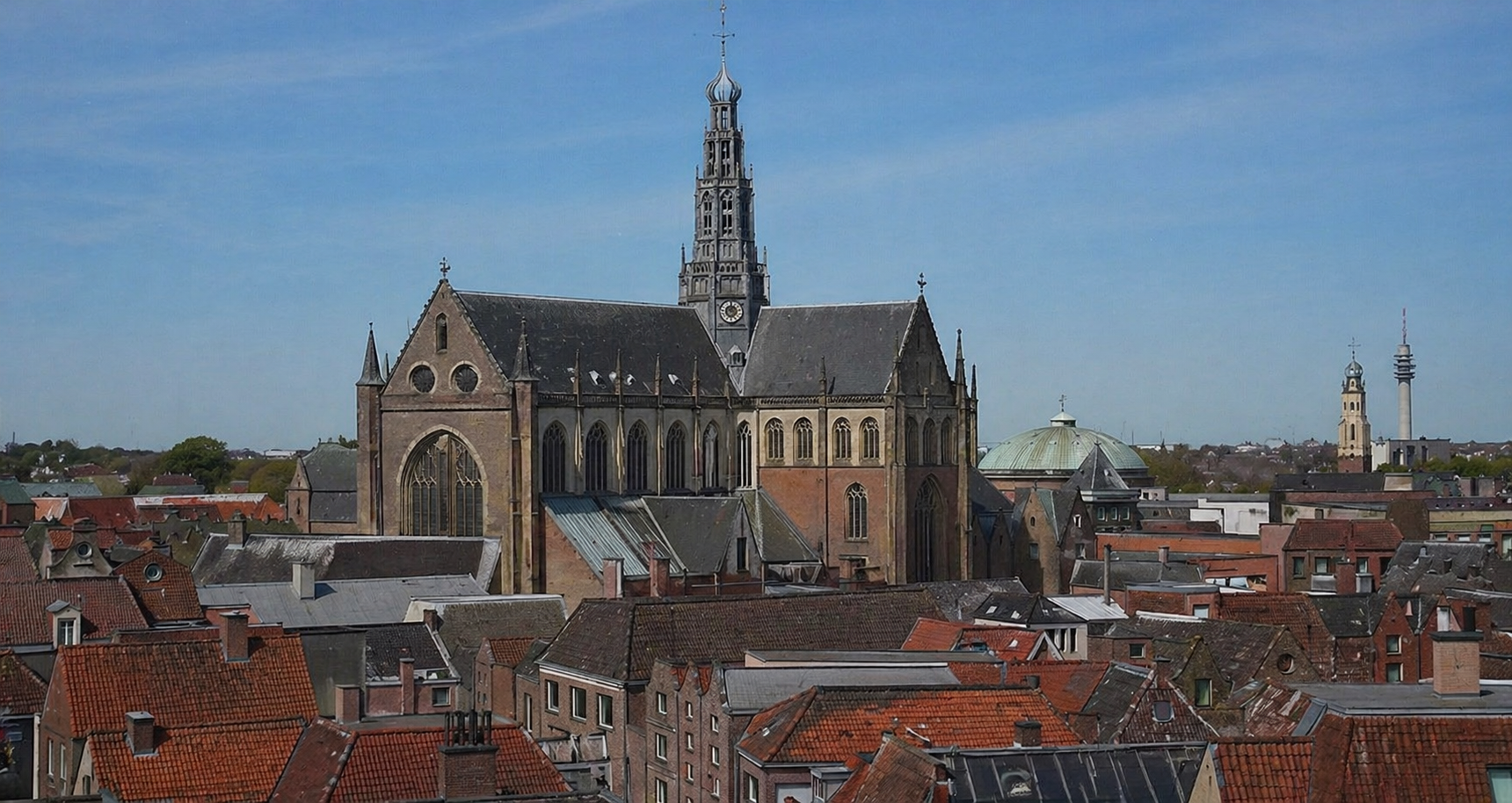 Uitzicht over stadsgedeelte met een grote gotische kerk als centraal punt en rode dakpannen op omliggende gebouwen bij een blauwe lucht.