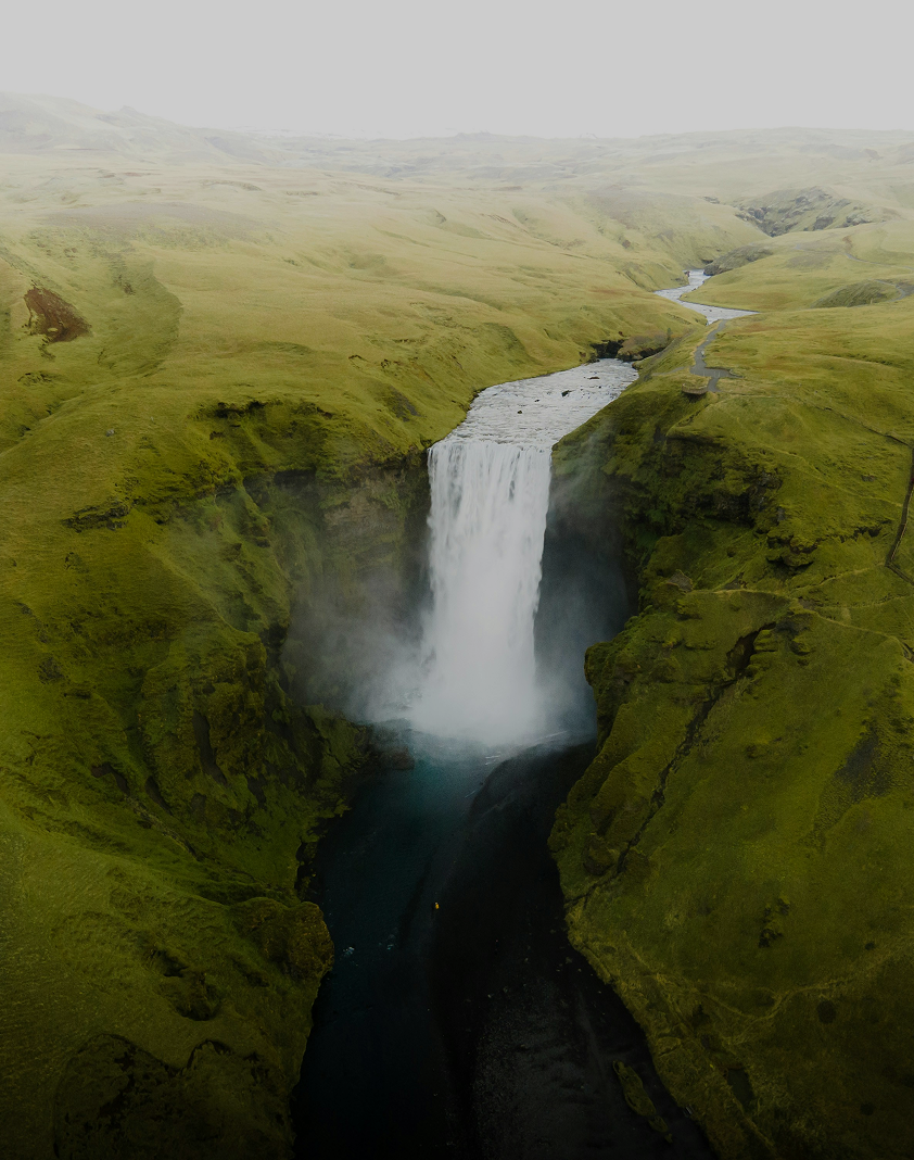 Tall waterfall flowing between moss-covered cliffs into a dark river below under a misty sky.
