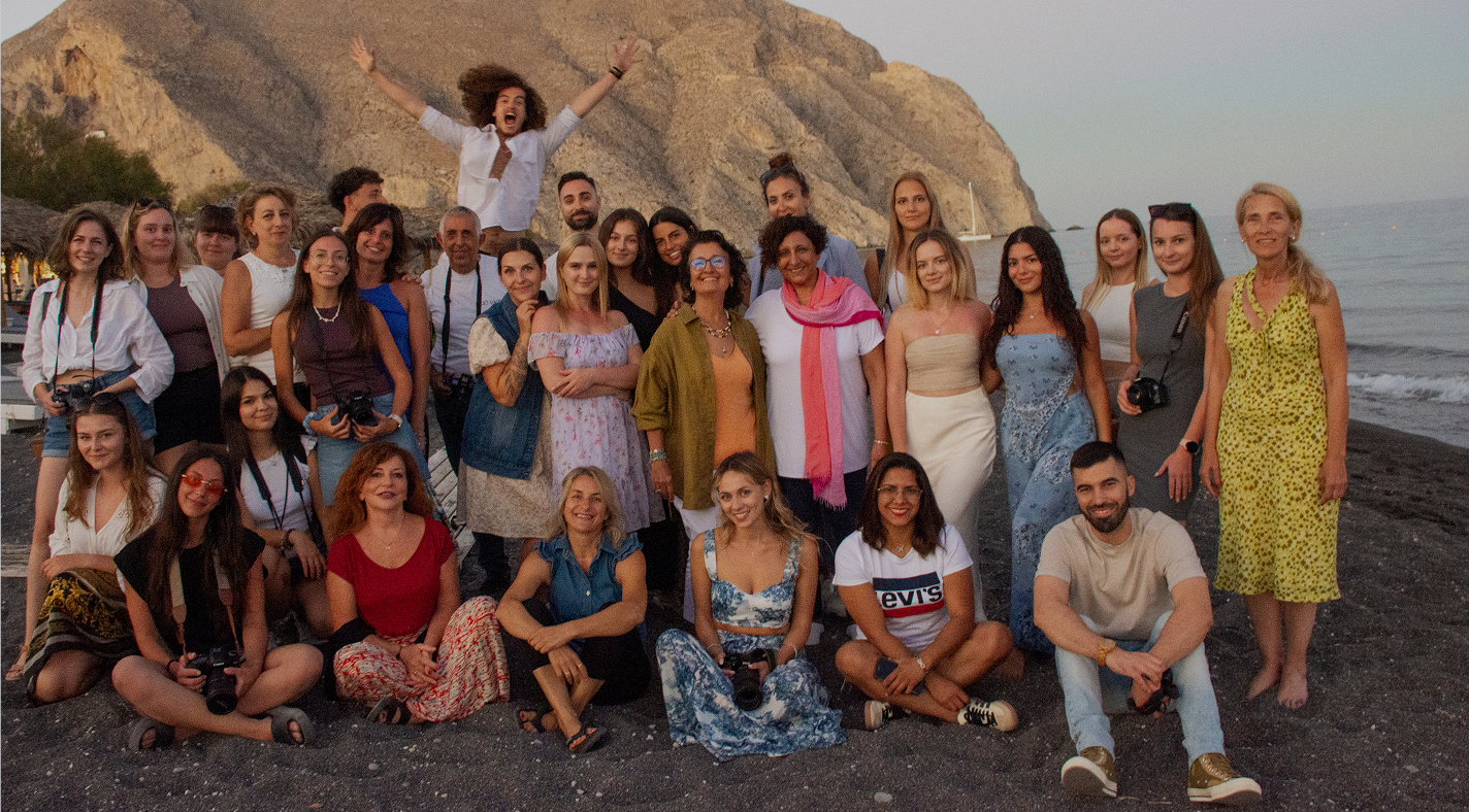 Group of diverse people posing and smiling at the camera on a beach with rocky cliffs and sea in the background at sunset.