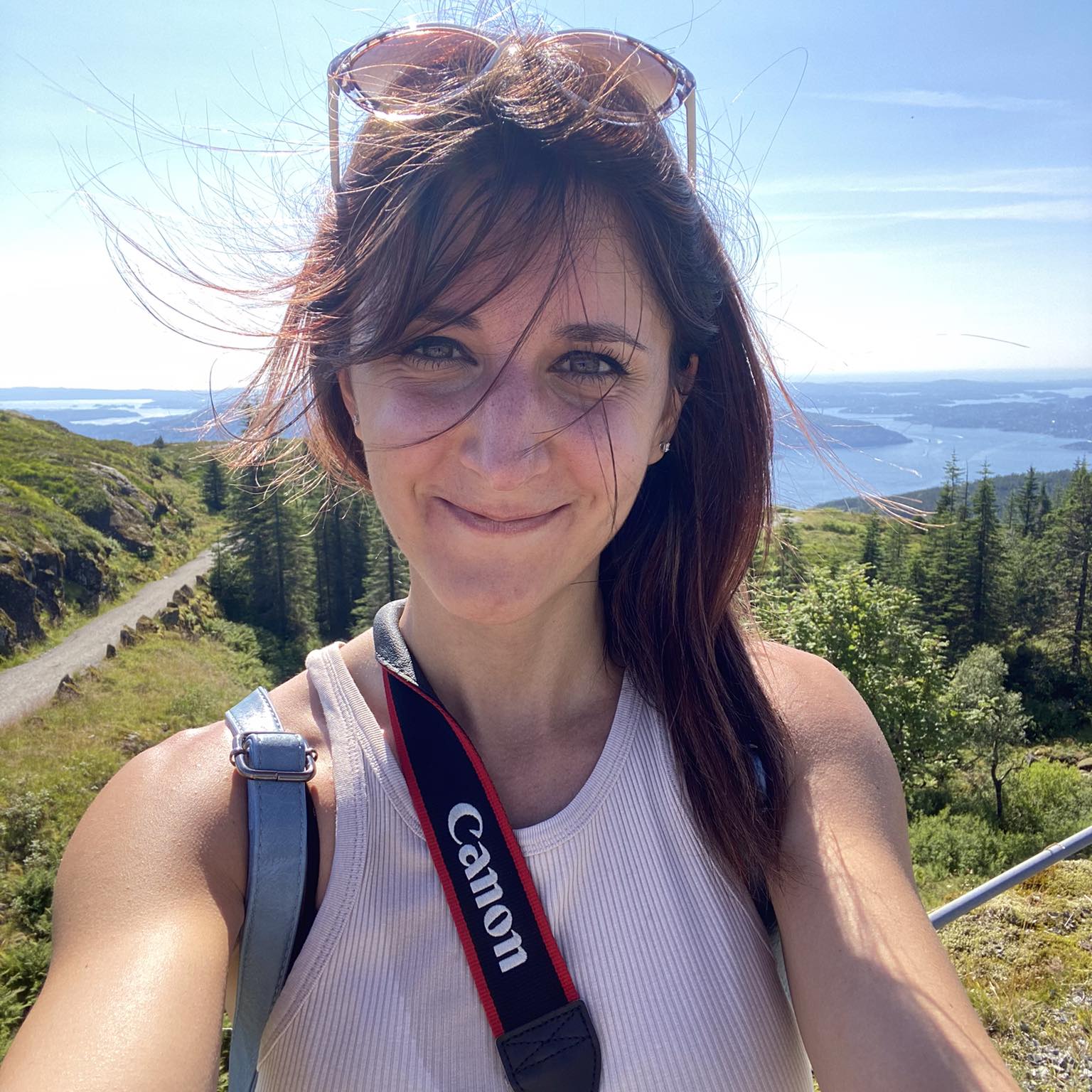 Smiling woman with sunglasses on her head and a Canon camera strap around her neck standing outdoors on a sunny day with green hills and a view of water and islands in the background.
