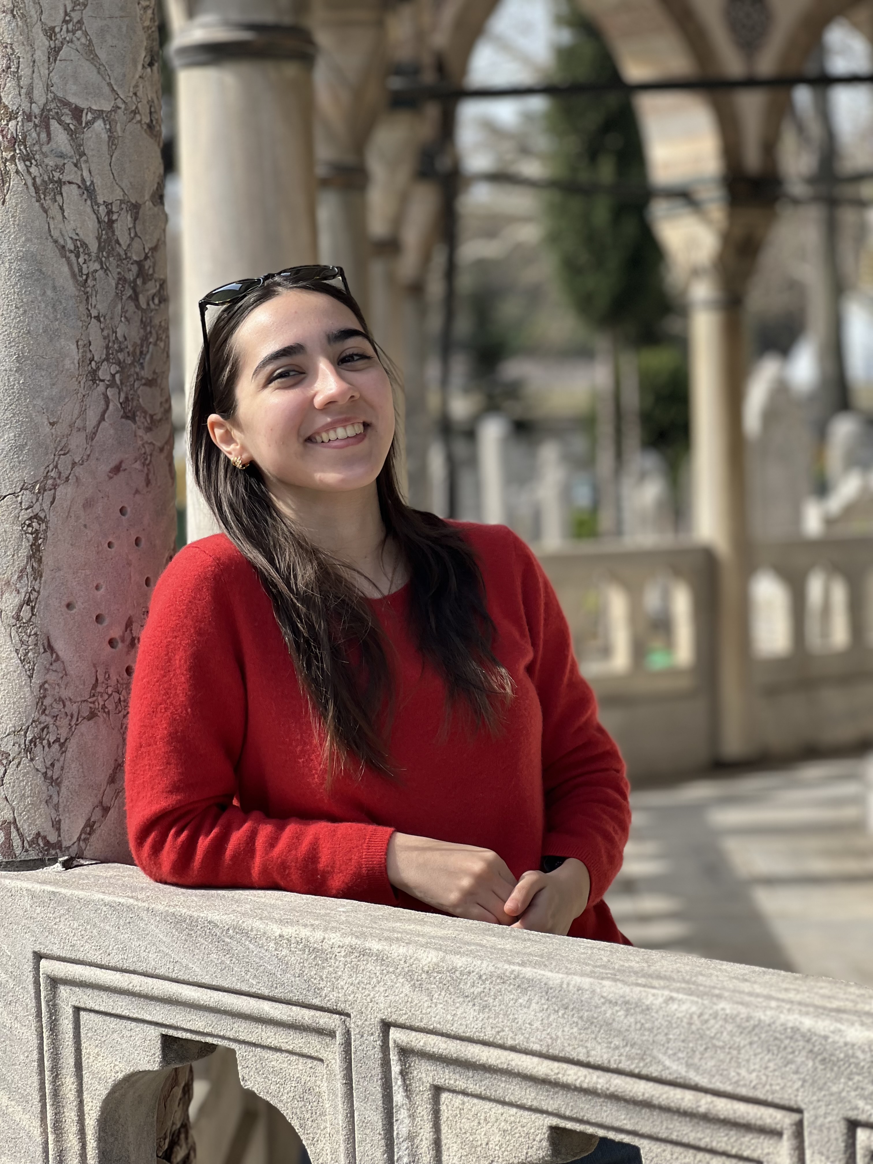 Smiling woman in a red sweater leaning on a stone railing with marble columns and arches in the background.
