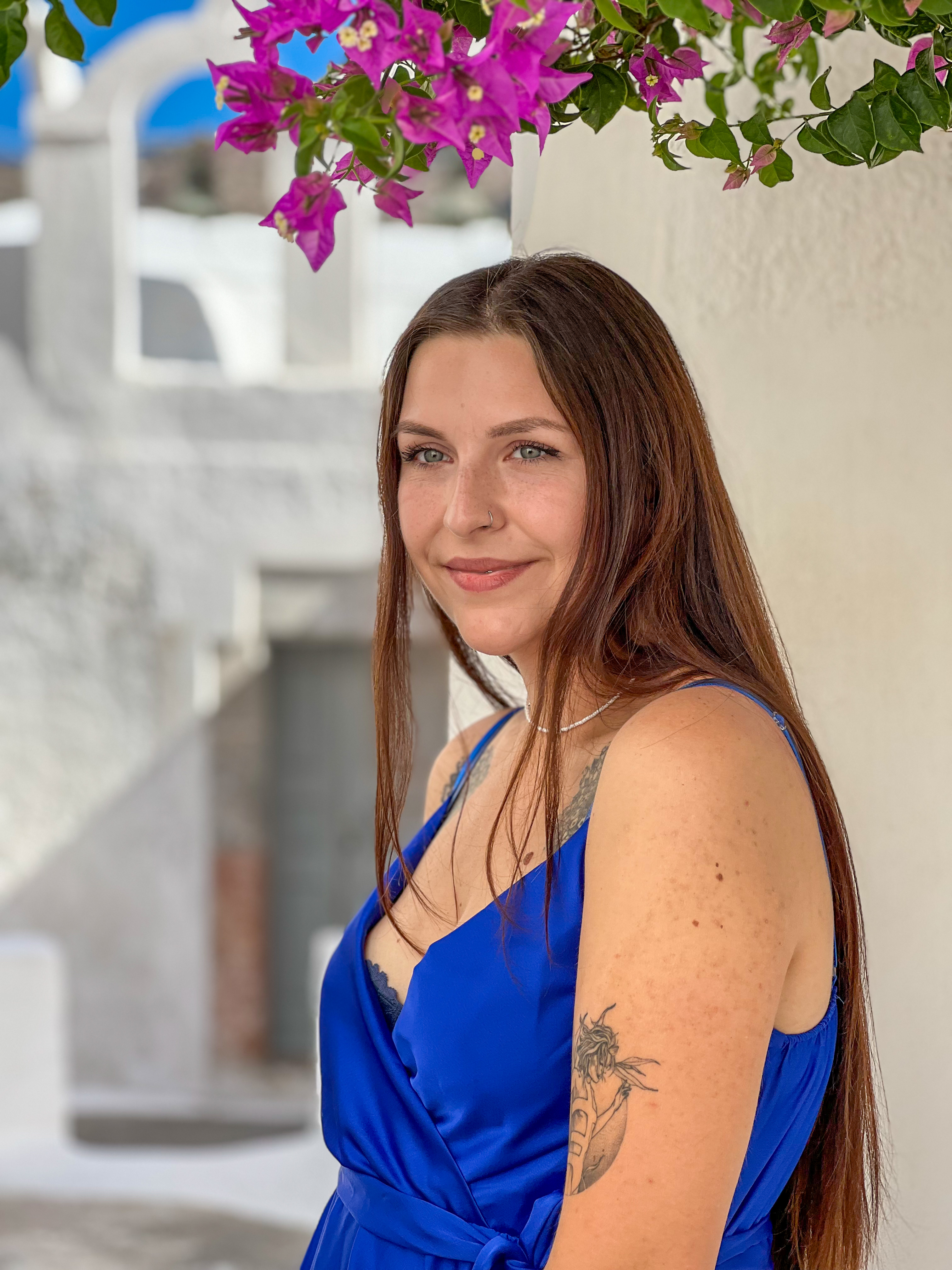 Young woman with long brown hair wearing a blue dress and a tattoo on her upper arm, standing against a white wall with purple flowers above.