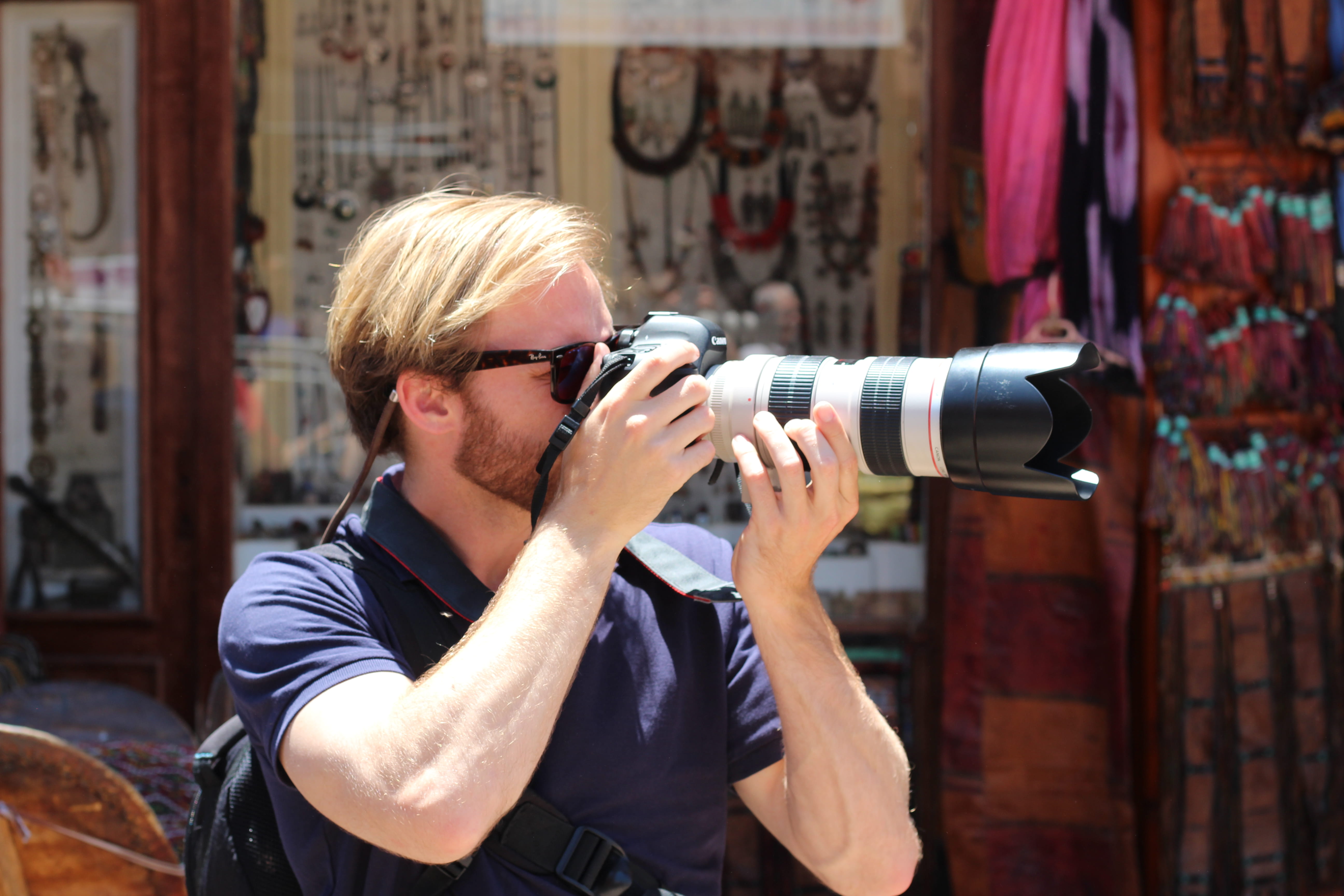 Blonde man wearing sunglasses and a navy blue shirt, holding a Canon camera with a large telephoto lens, taking a photo outdoors.