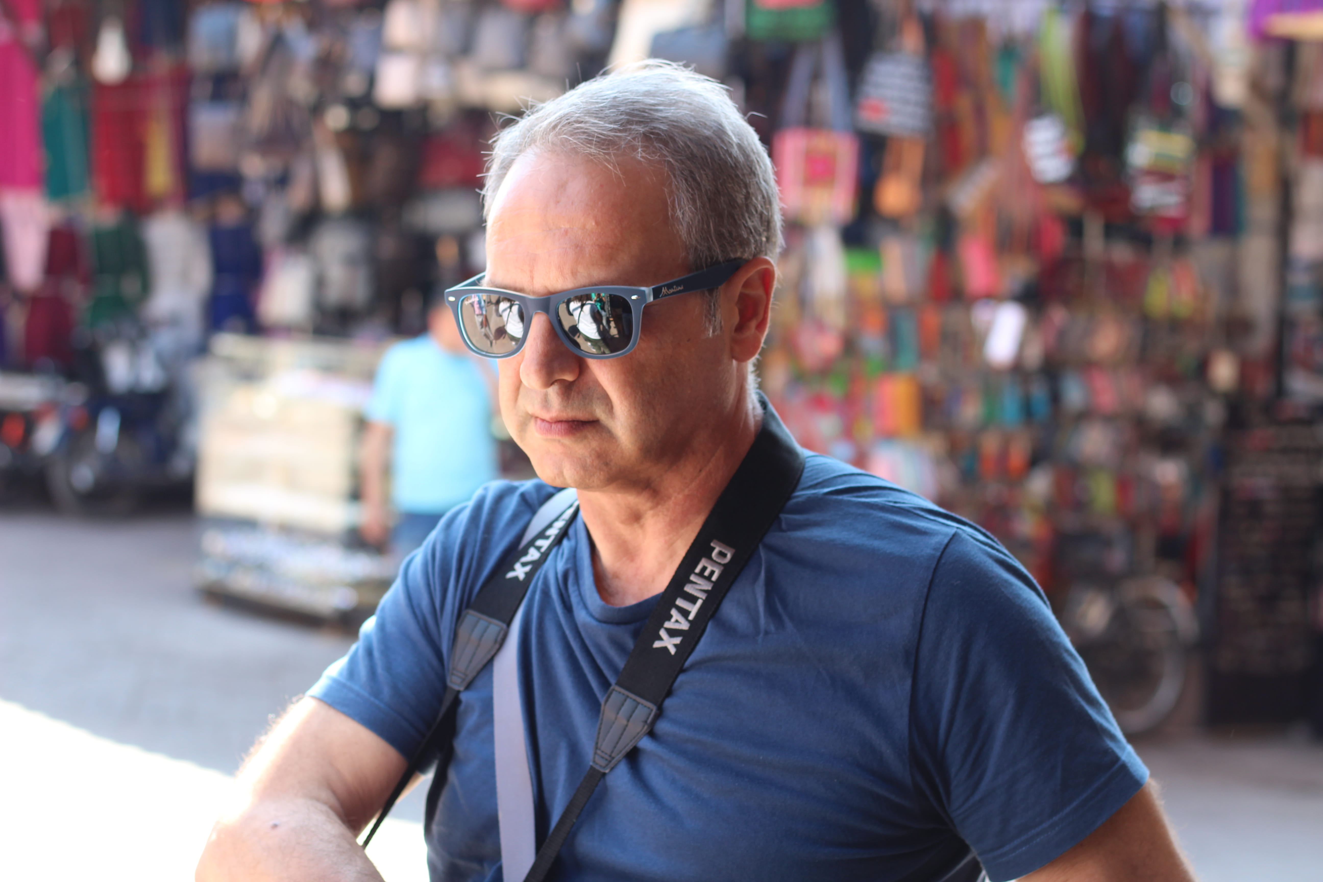 Middle-aged man with grey hair wearing blue sunglasses and a blue t-shirt with a camera strap around his neck in a market setting.