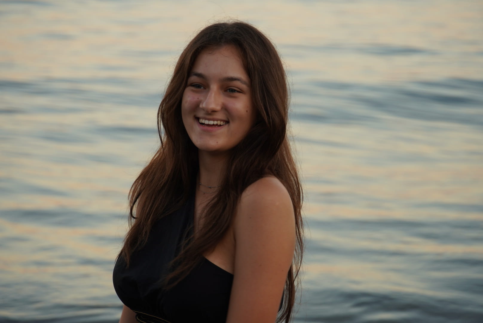 Young woman with long brown hair smiling by calm water during sunset.