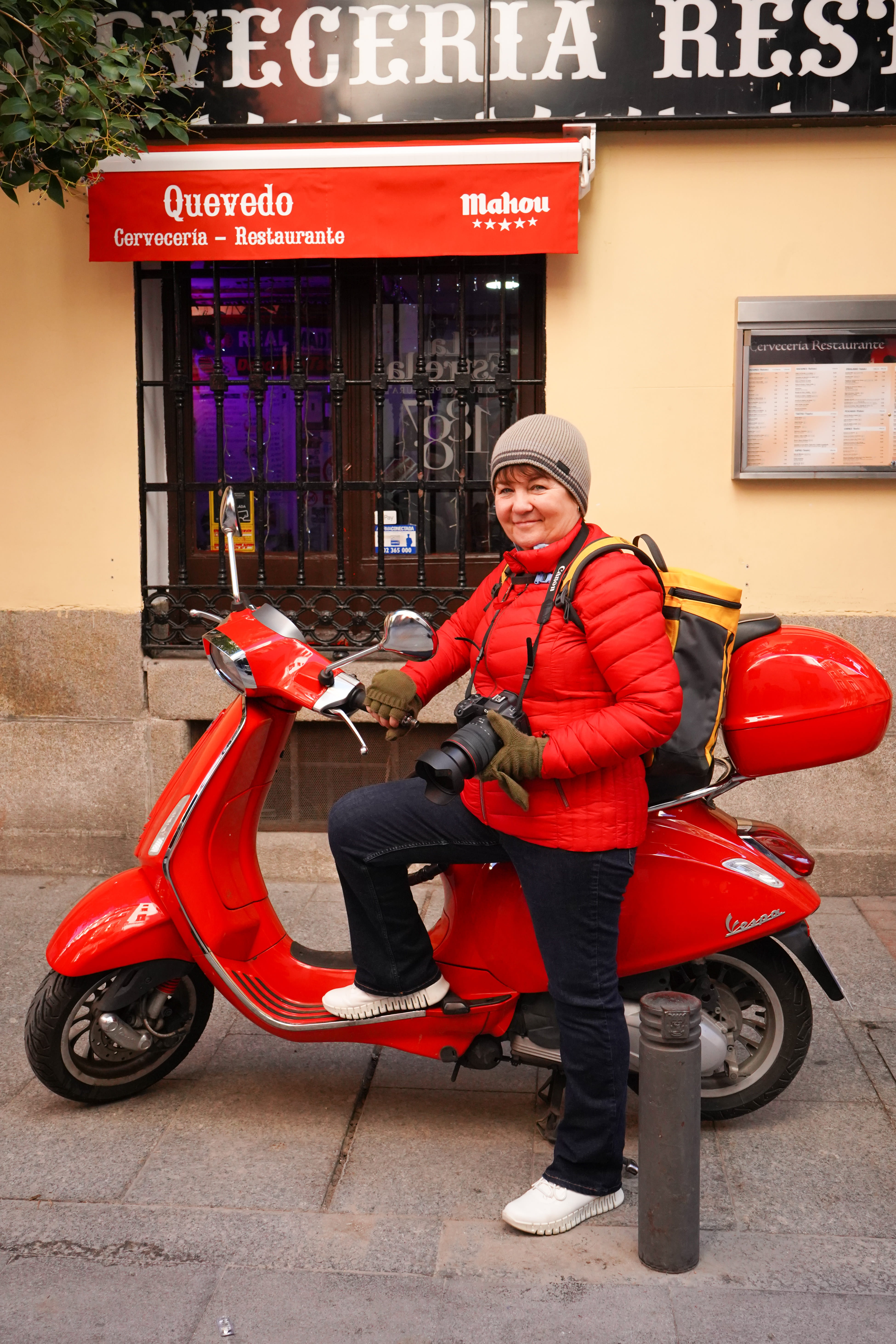 Woman in red jacket and grey beanie holding a camera while standing beside a red Vespa scooter outside a restaurant.