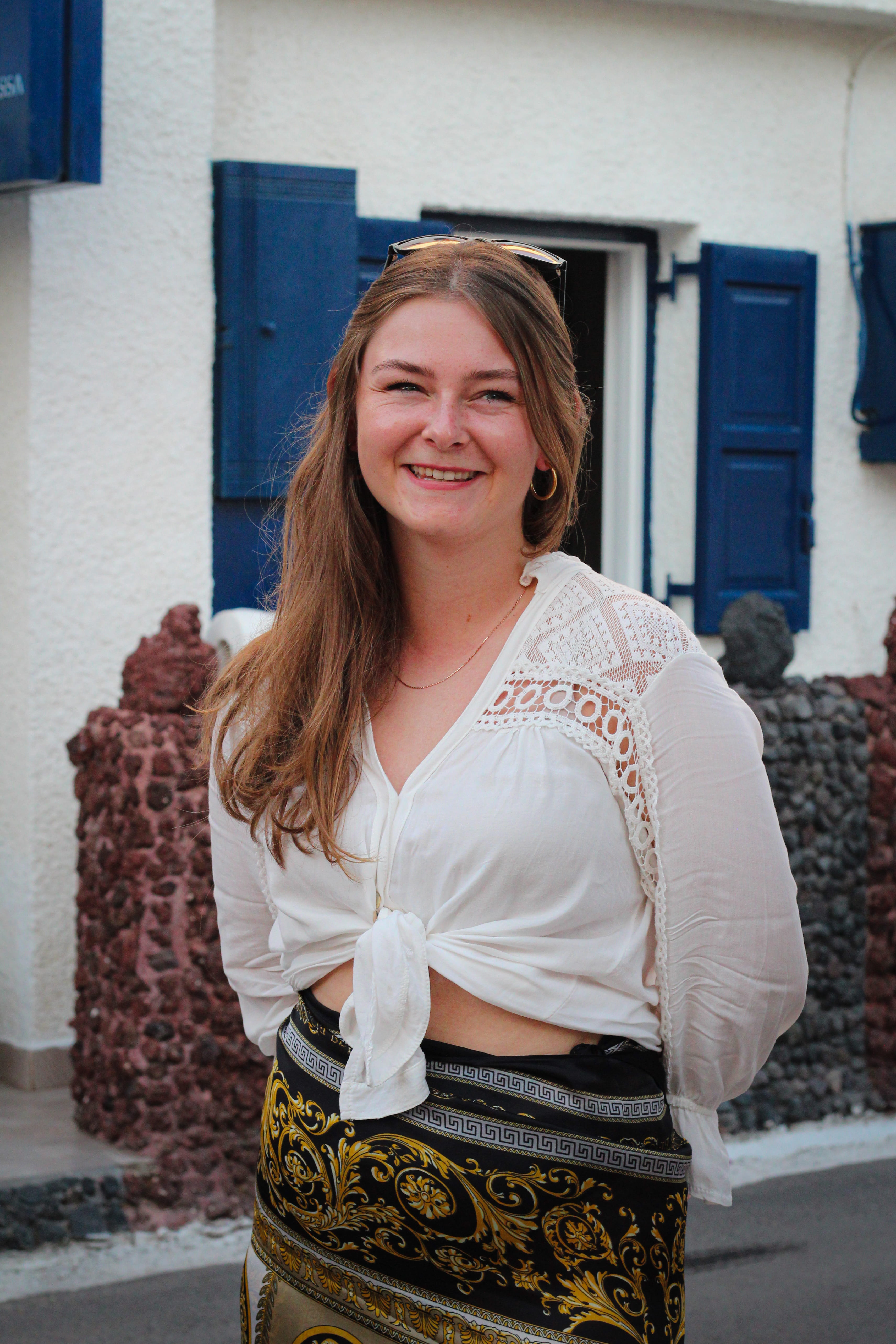 Smiling woman with long hair wearing a white tied blouse and patterned skirt standing outdoors near a white building with blue shutters.