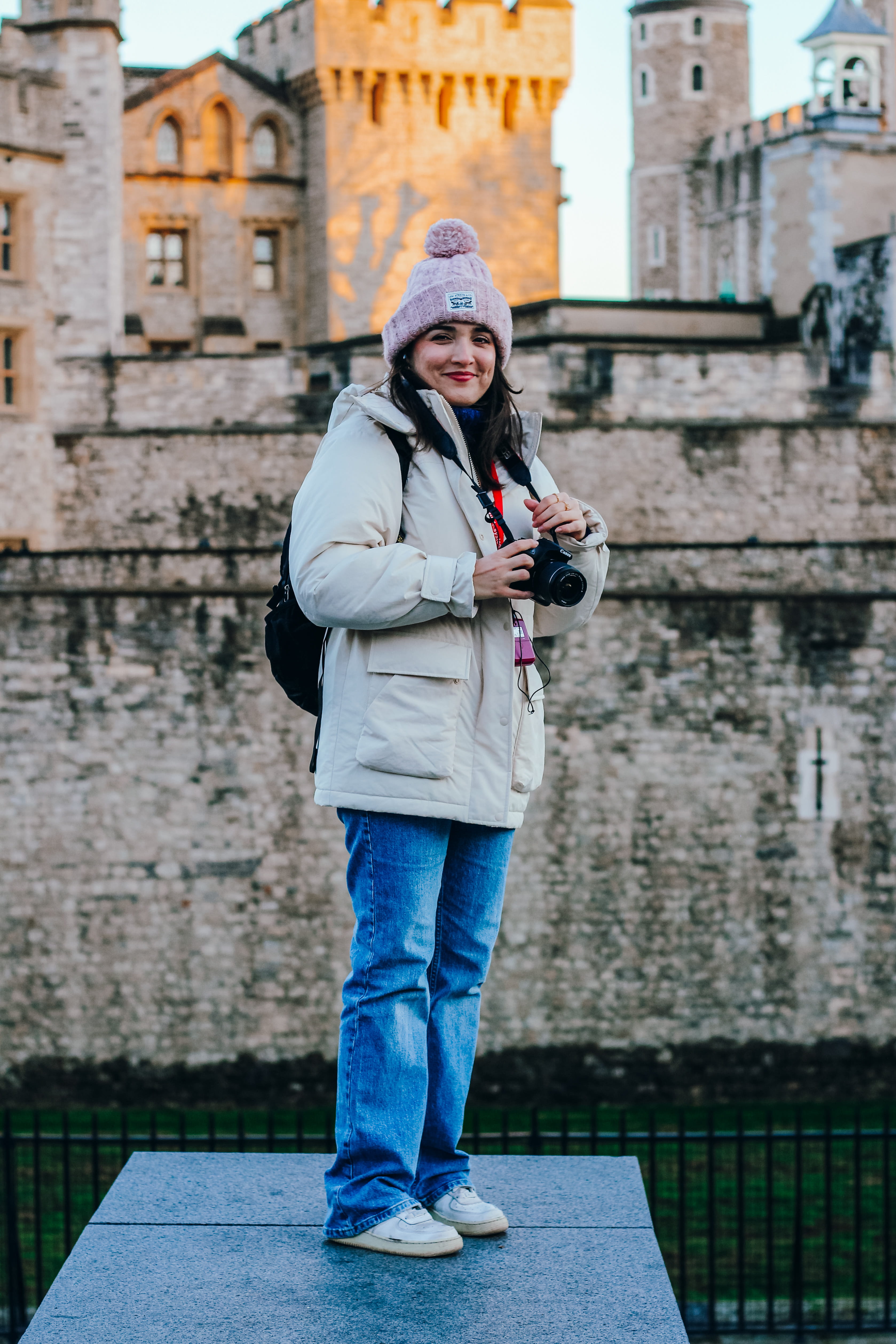 Woman in a beige coat and pink beanie holding a camera standing in front of a historic stone castle.
