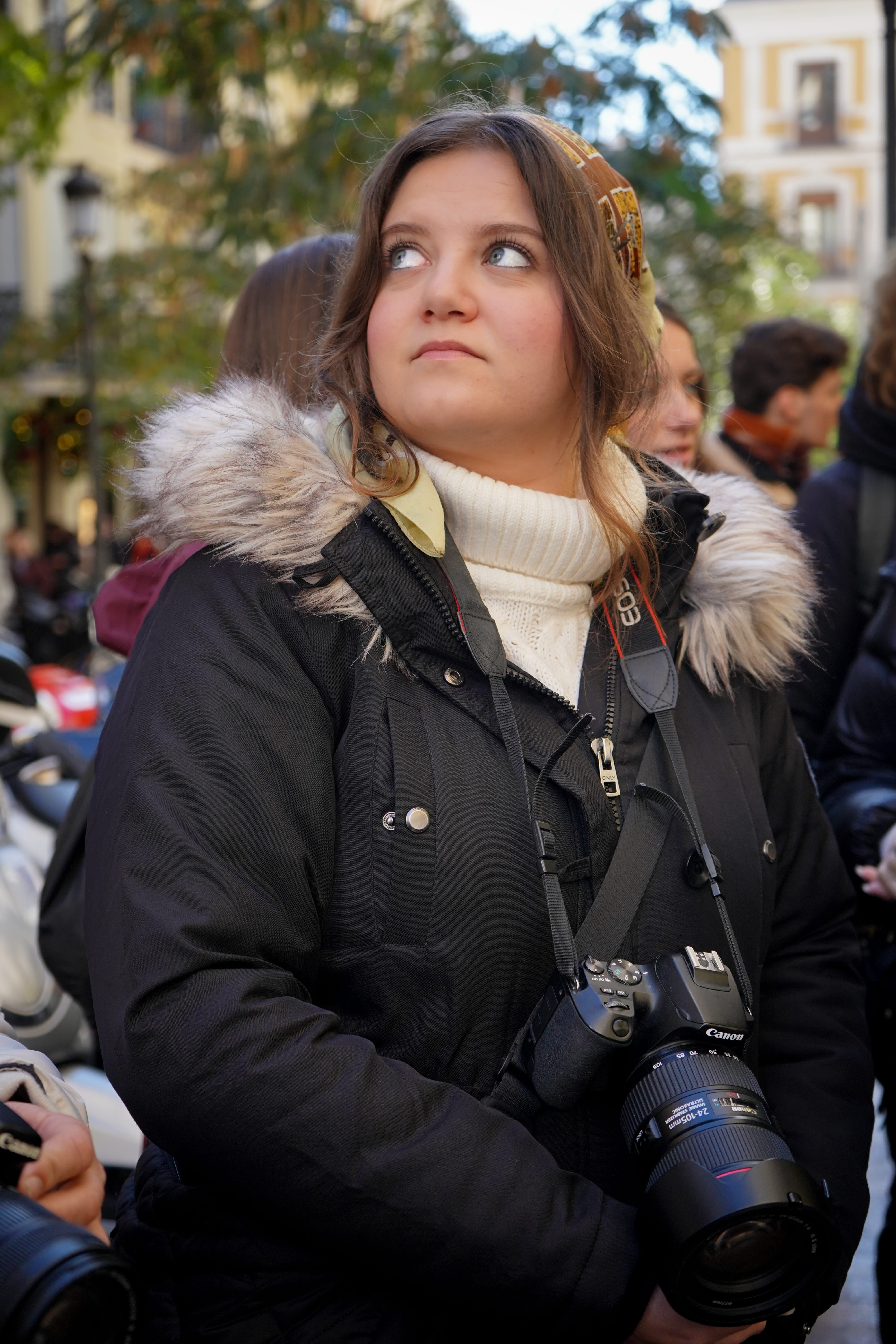 Young woman in a black coat with fur collar holding a Canon camera, looking up and to the side outdoors.