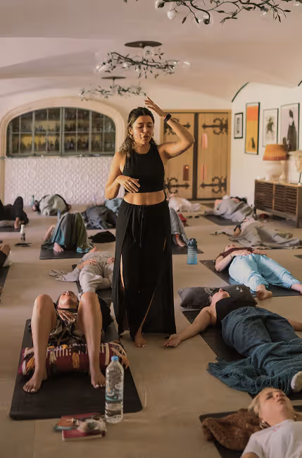 Yoga instructor leading a class with participants lying on mats in a spacious, softly lit room.
