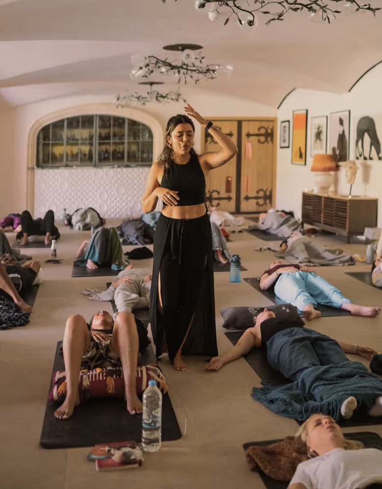 Melanie Lopes standing and guiding a group lying on mats in a calm, softly lit studio with various artworks on the walls.