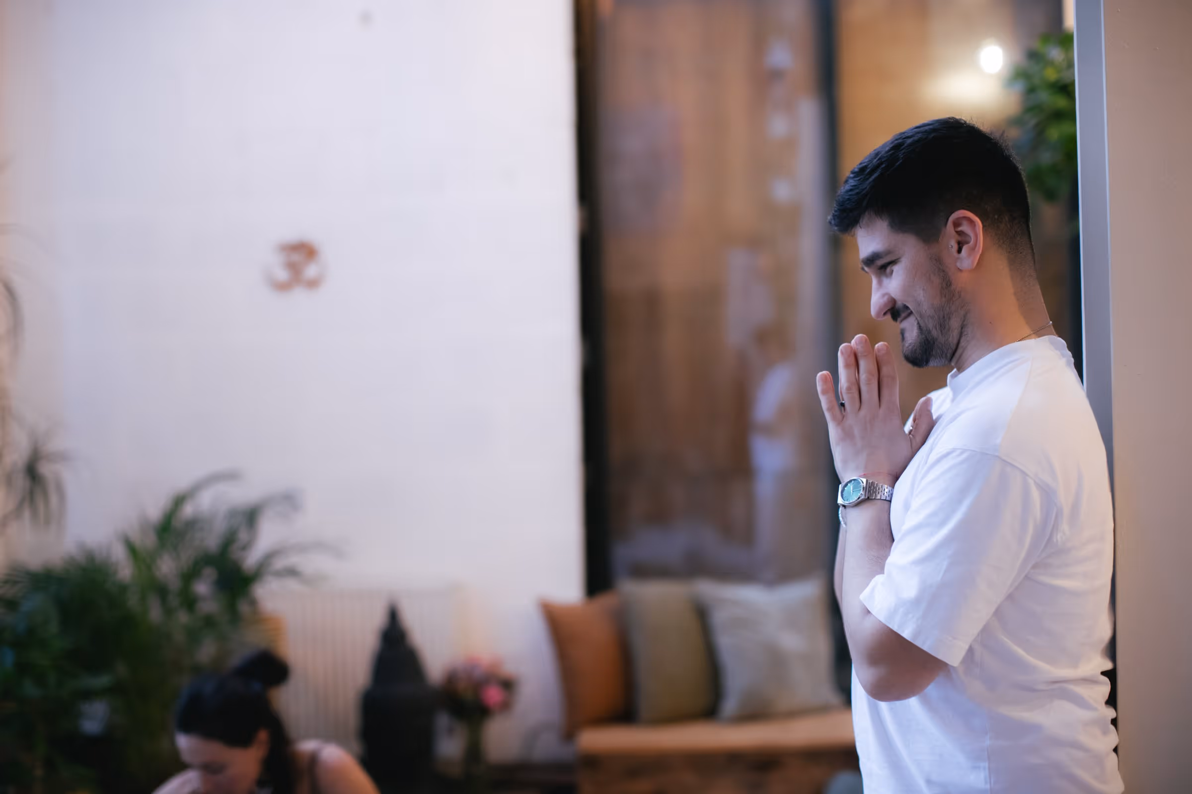 Man in white shirt standing with hands pressed together in a peaceful gesture indoors.