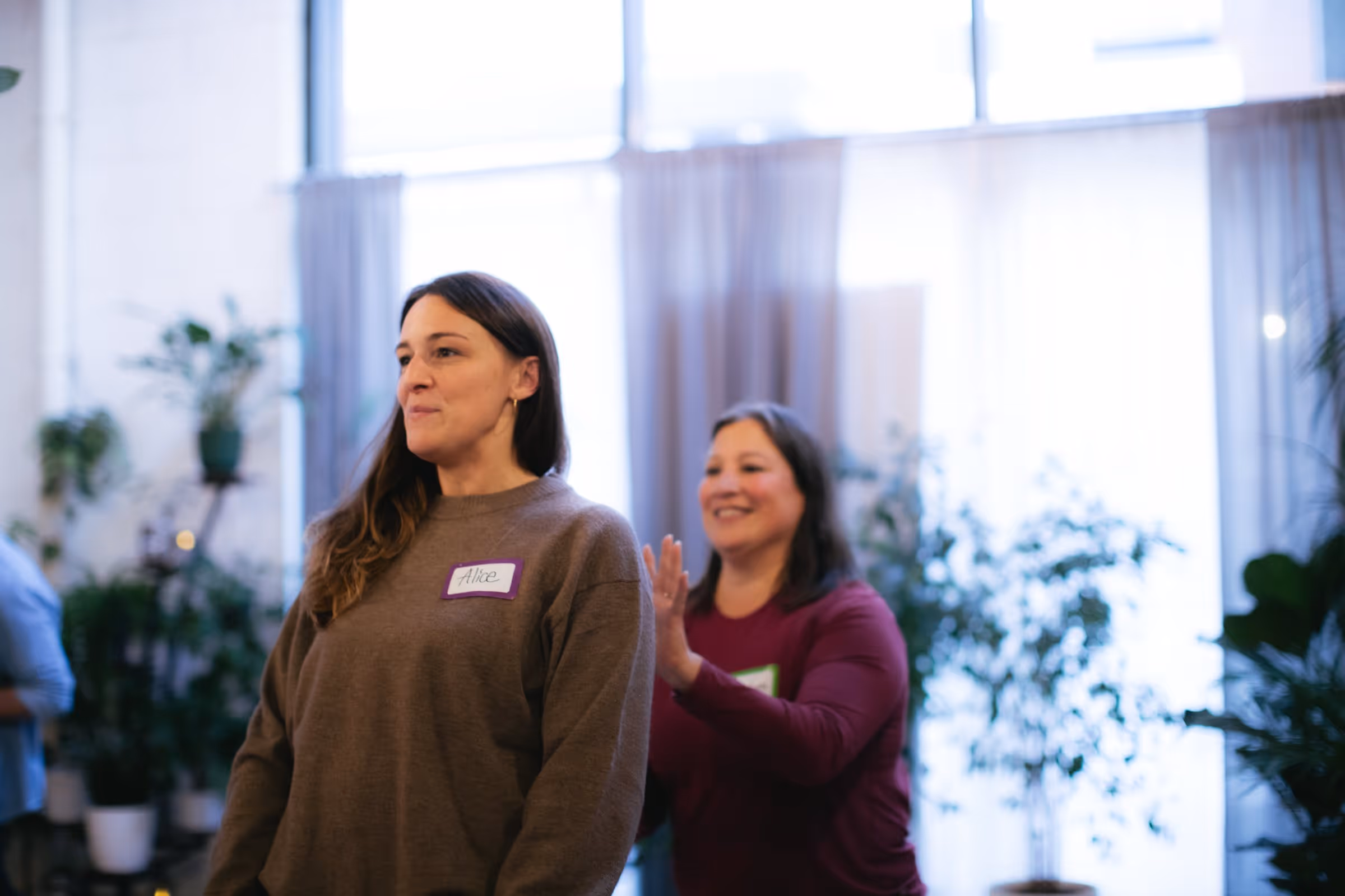 Two women standing indoors near plants, one smiling and reaching out toward the other wearing a name tag 'Alice'.