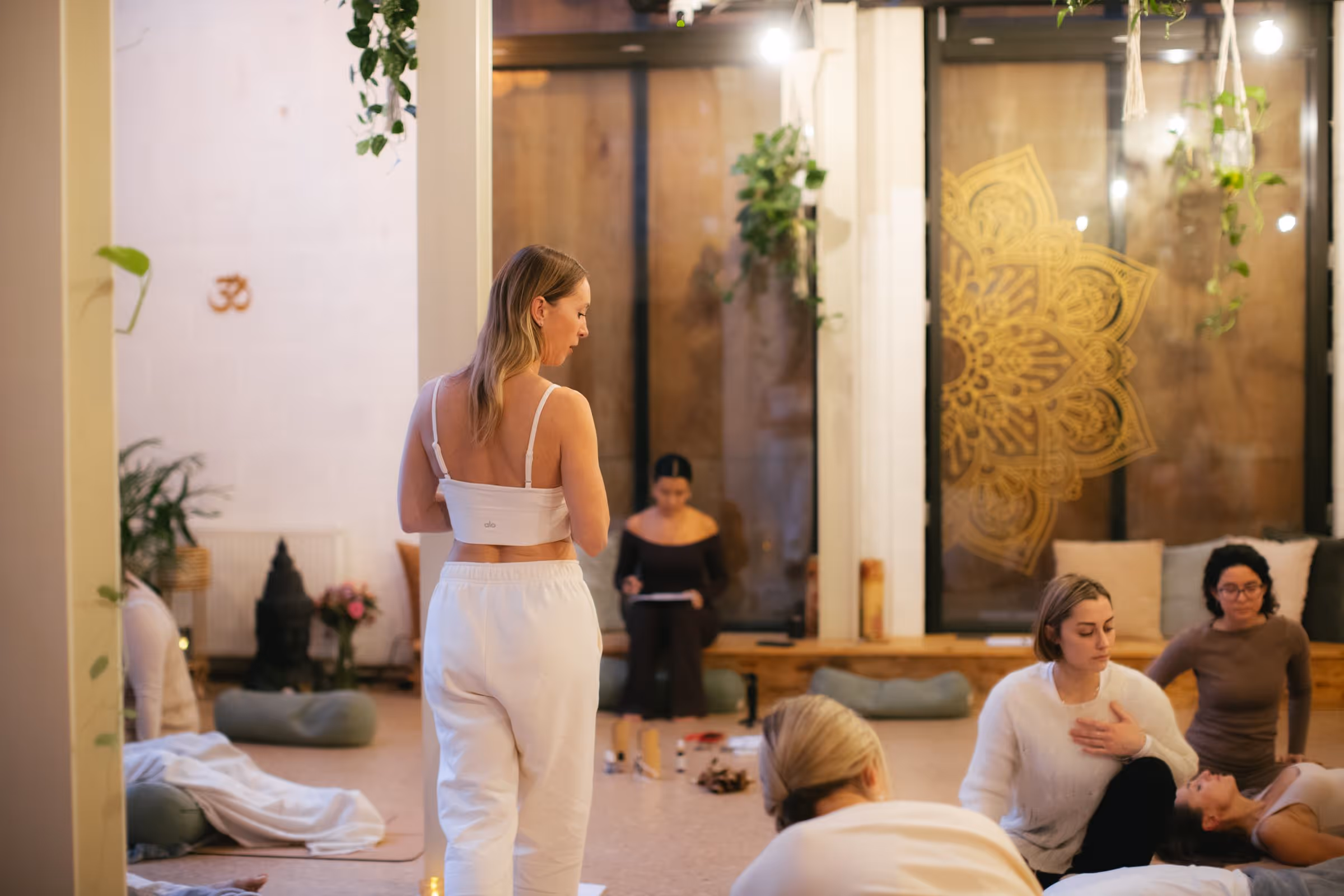 Woman in white outfit standing and overseeing a group session in a peaceful, plant-decorated room.