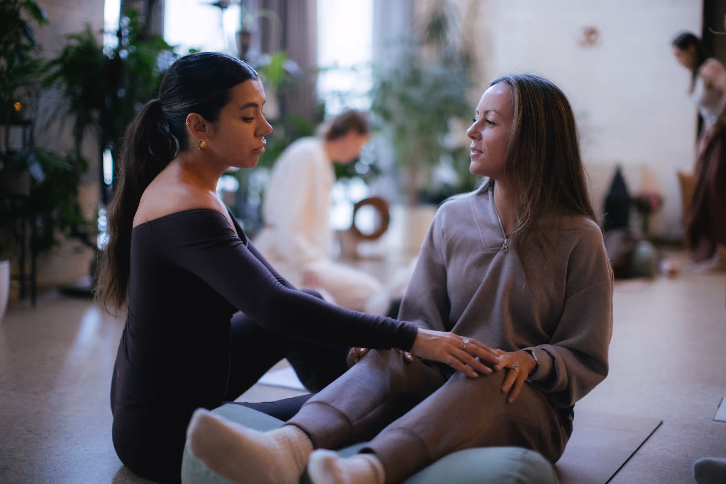 Two women sitting on floor cushions facing each other, one gently holding the other's knee in a calm indoor setting.