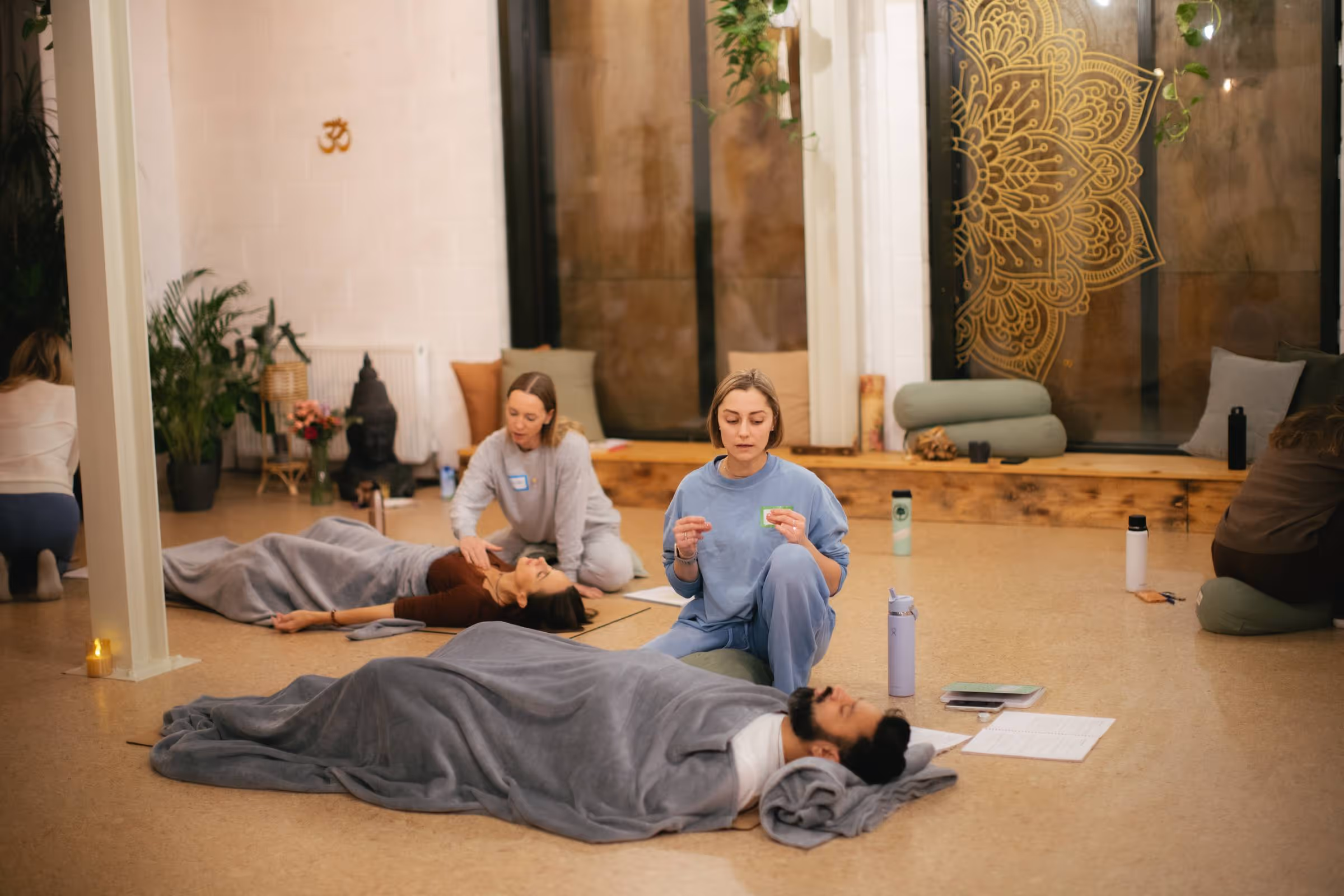Group session in a meditation room with people lying under blankets and others seated near them.