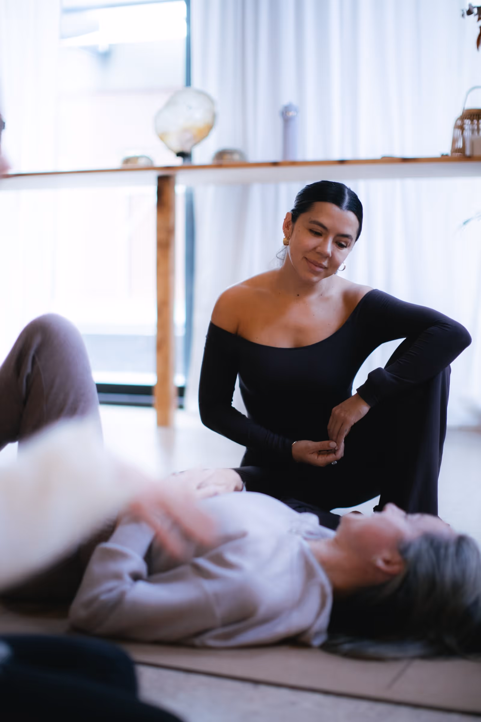Woman in black sitting on the floor attentively listening to a person lying down in a bright room.