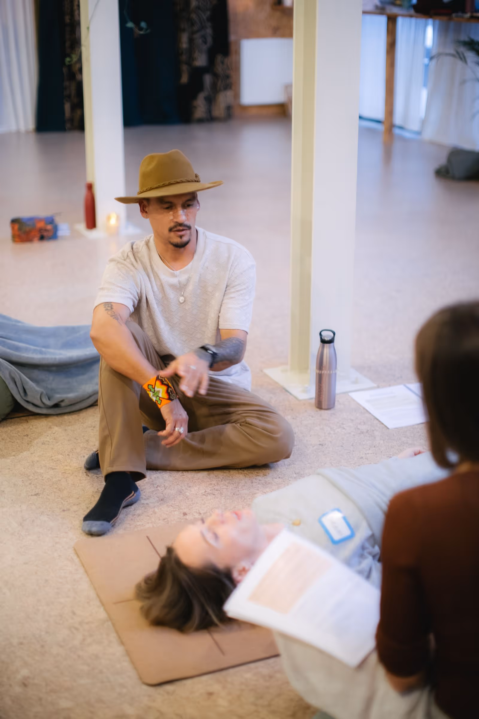 Man wearing a brown hat and beige shirt sitting cross-legged on the floor explaining breathwork to a reclining woman.
