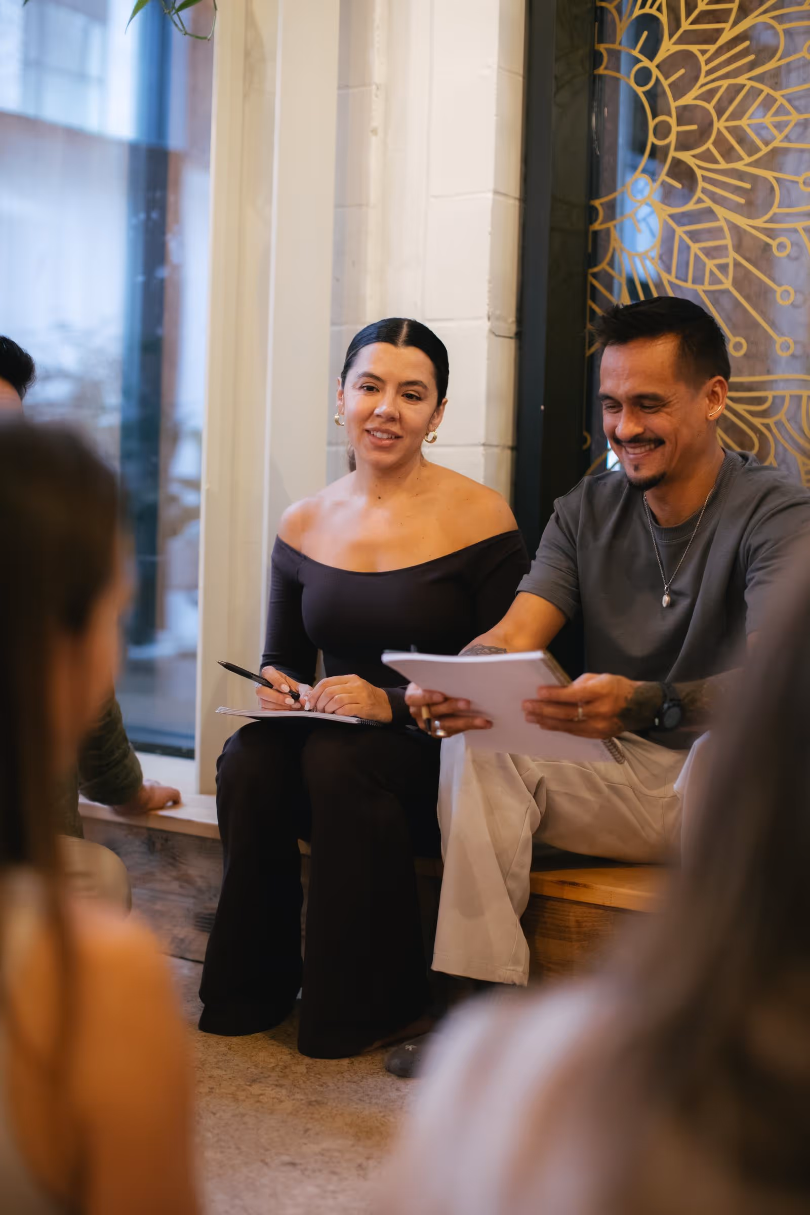Two adults sitting indoors, smiling and holding notebooks, engaged in a discussion.