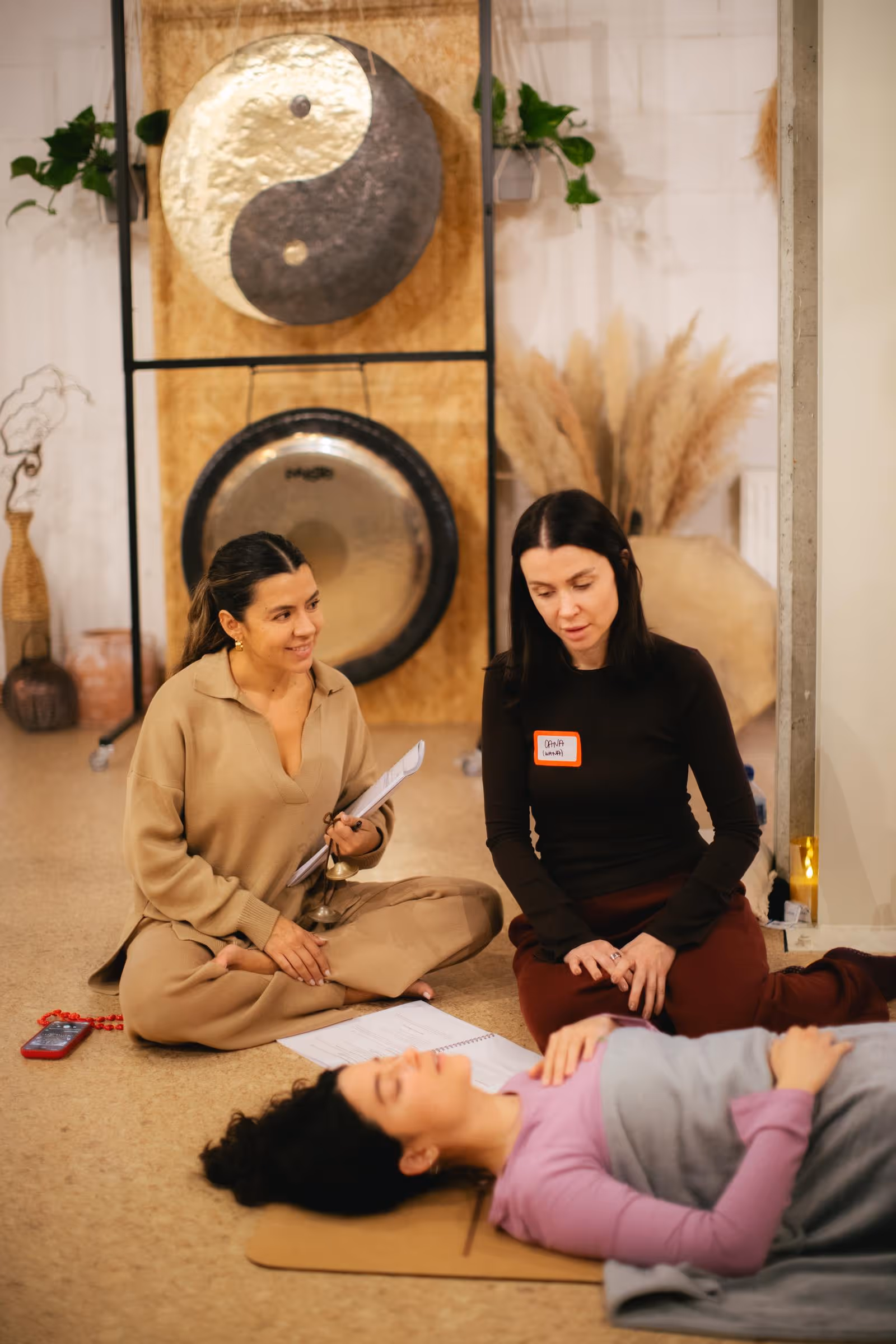 Two women sitting cross-legged on the floor near a yin-yang gong, another woman lies relaxed on a mat covered with a blanket.