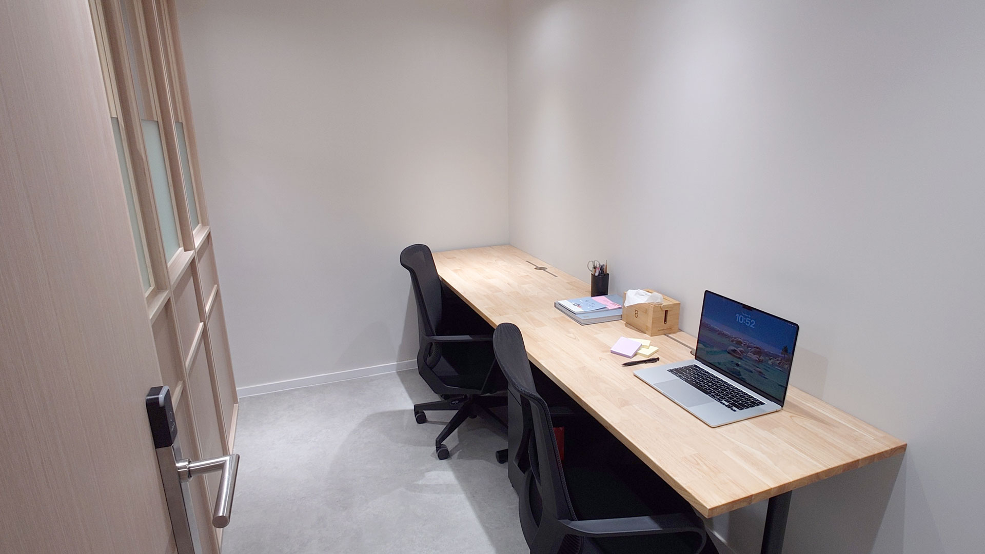Minimalist office room with light wood door left ajar, light wood desk along the right wall holding a laptop, books, tissue box, pens, sticky notes, and two black office chairs on a light gray floor.