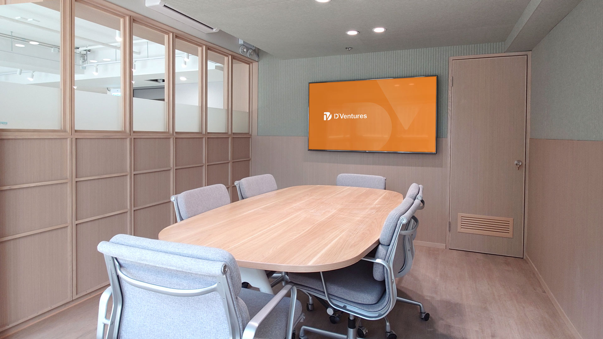 Modern conference room with a light wooden oval table surrounded by six gray ergonomic chairs on casters, light wood-paneled walls, a closed door, and a large flat screen displaying an orange slide with the text 'D'Ventures'.