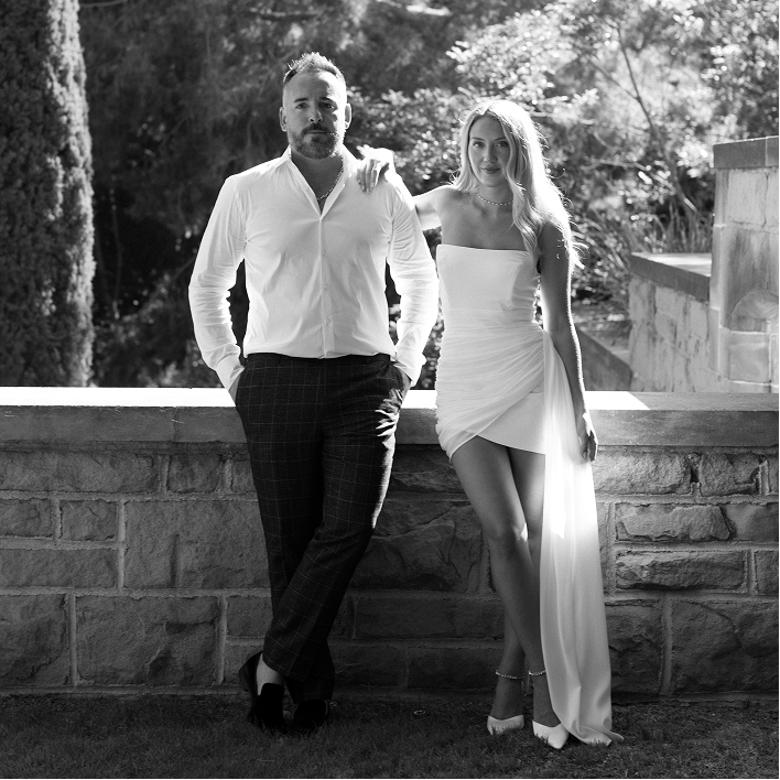 Black and white photo of man in white shirt and woman in white dress leaning on stone wall outdoors.
