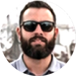 Bearded man wearing dark sunglasses and a collared shirt with a blurred outdoor background.
