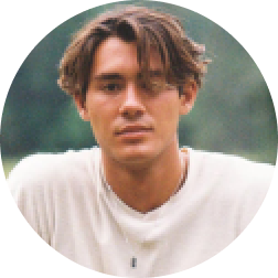 Young man with medium-length brown hair wearing a white shirt against a blurred outdoor background.