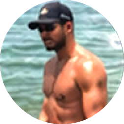 Shirtless man wearing sunglasses and a cap standing near water.