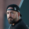 Bearded man with backward black cap in a dark shirt against a dark blue background.