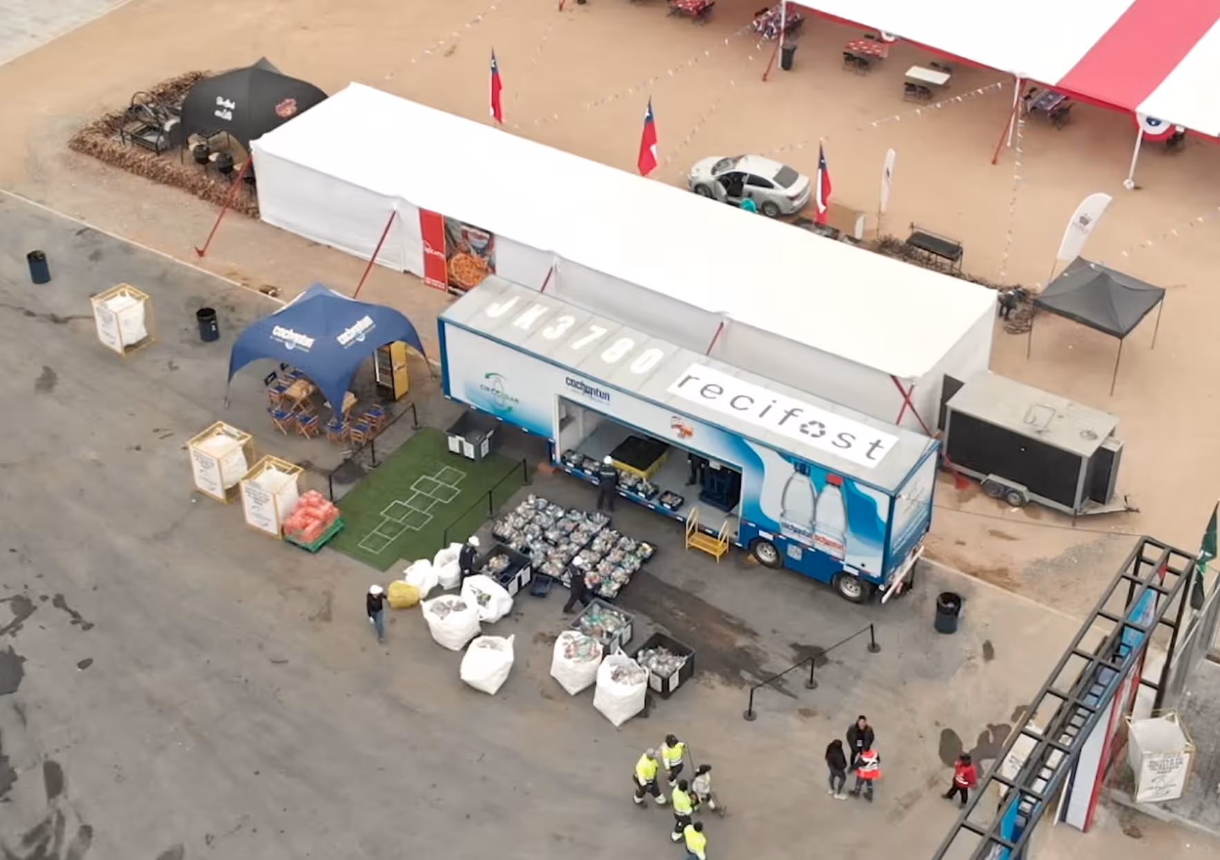 Vista aérea de un camión reciclador Recifest con bolsas y cajas llenas de materiales reciclables, carpas y personas en un evento al aire libre.