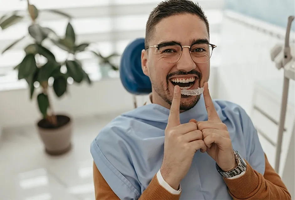 Man sitting in a dental chair smiling while holding a clear dental aligner in front of his teeth.