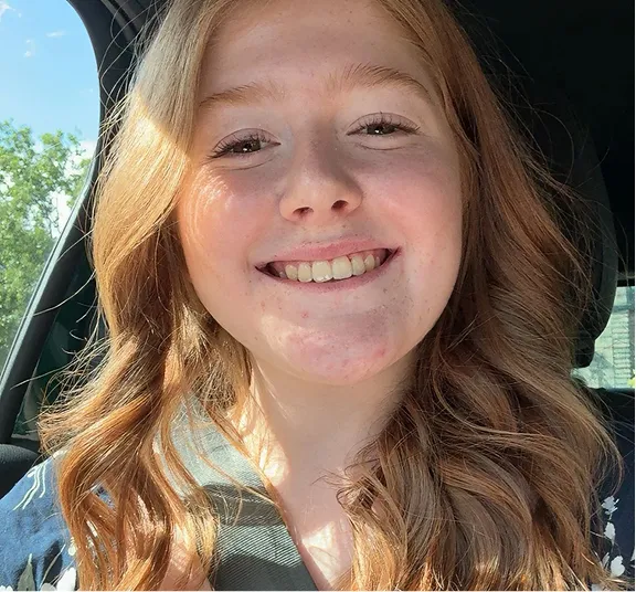 Smiling young woman with long wavy red hair sitting in a car wearing a seatbelt.