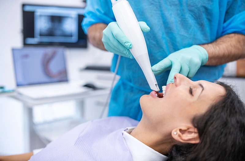 Dentist in gloves using a handheld scanner inside a woman's open mouth during a dental checkup.