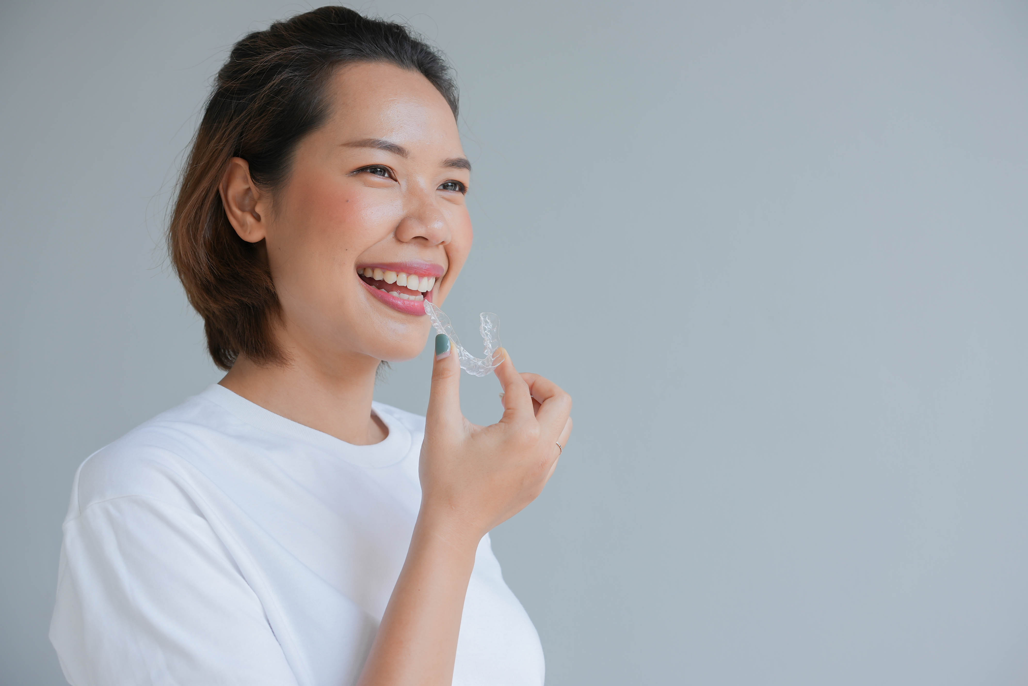 Smiling woman holding a clear dental aligner near her mouth against a plain gray background.