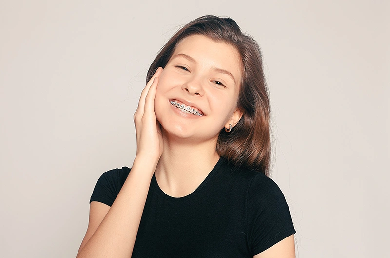 Smiling young woman with braces touching her cheek, wearing a black shirt against plain background.