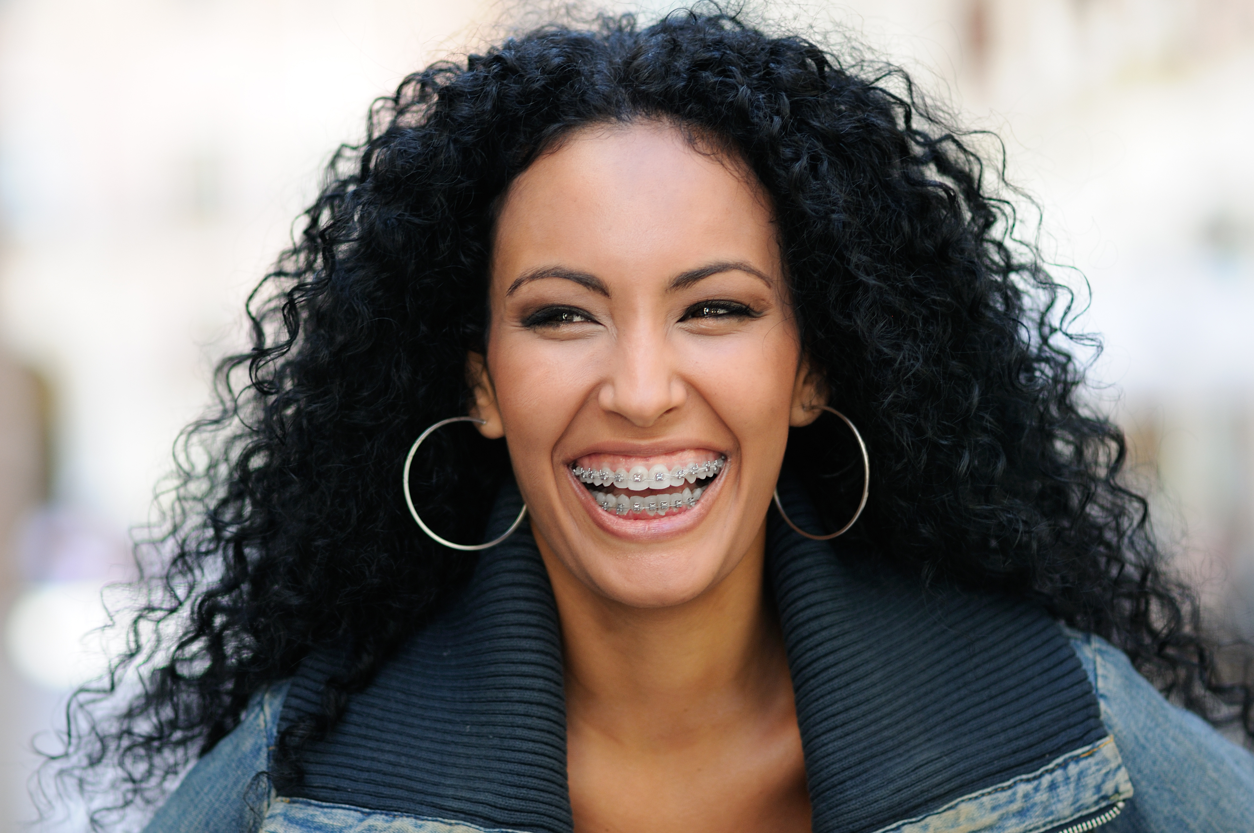 Woman with curly black hair and braces smiling brightly wearing large hoop earrings and a denim jacket.