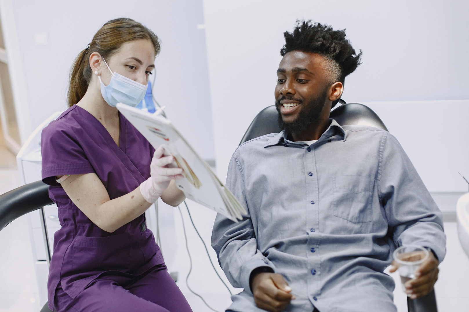 Dentist in purple scrubs showing a dental chart to a male patient holding a glass of water.