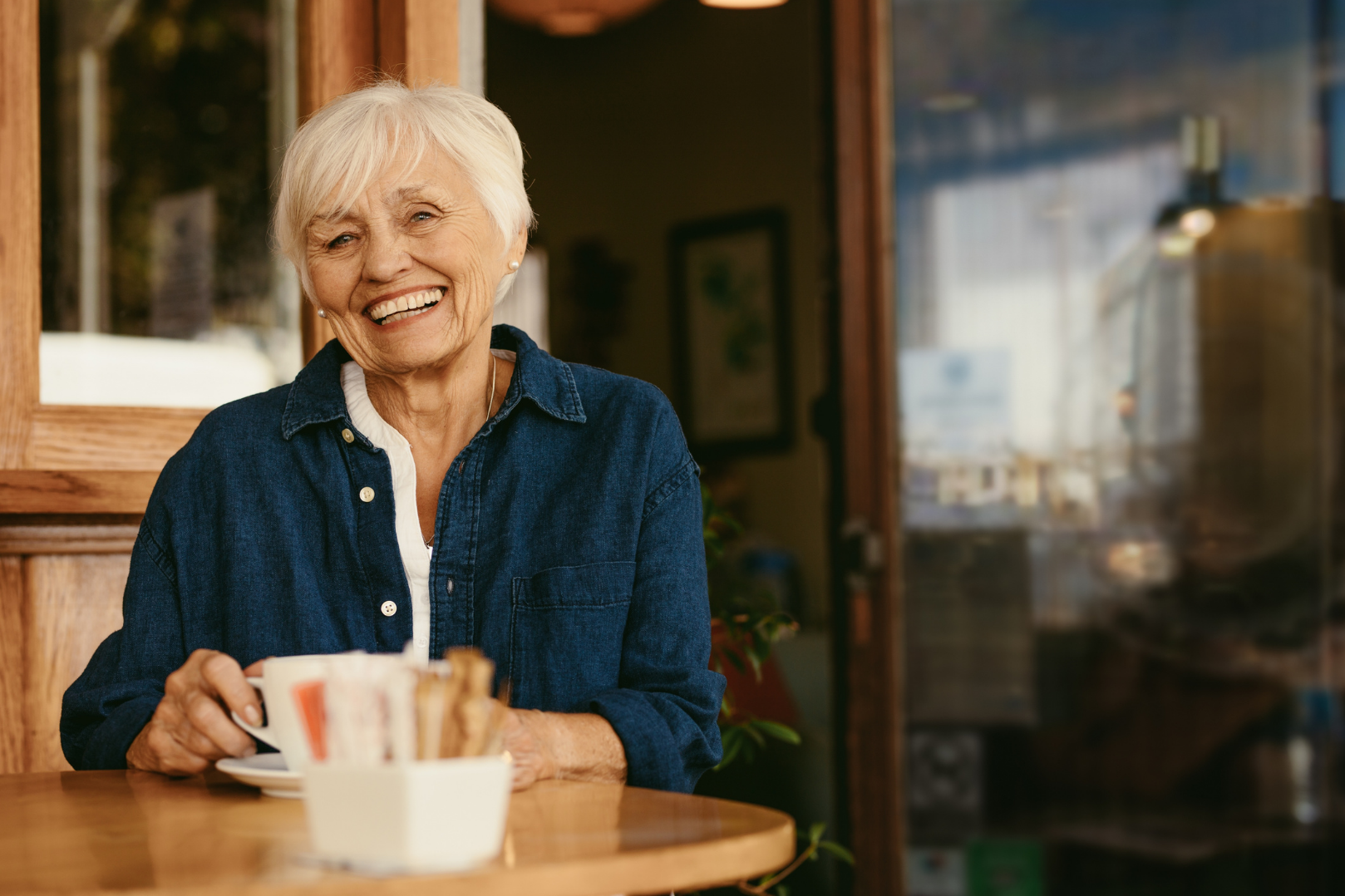 Smiling elderly woman in blue shirt holding a coffee cup at a wooden table in a cozy cafe.